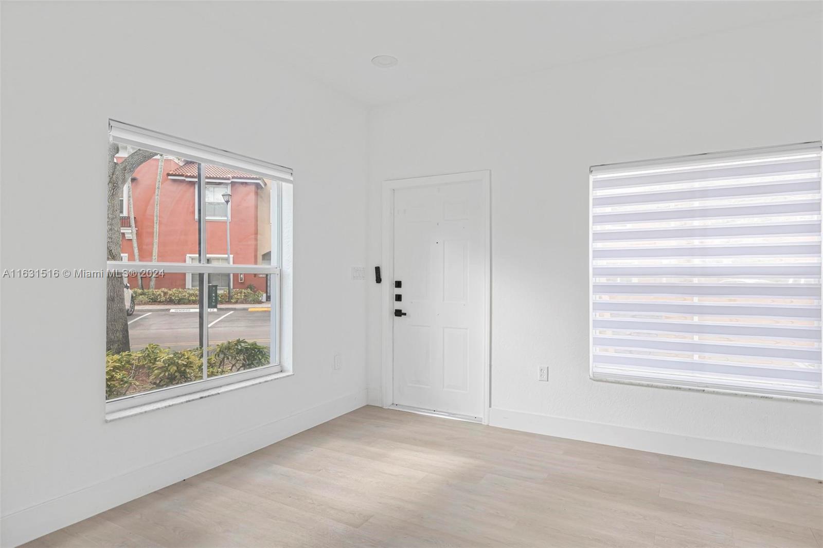 Vacant bright room with pale flooring, white entry door, and large windows with horizontal blinds.
