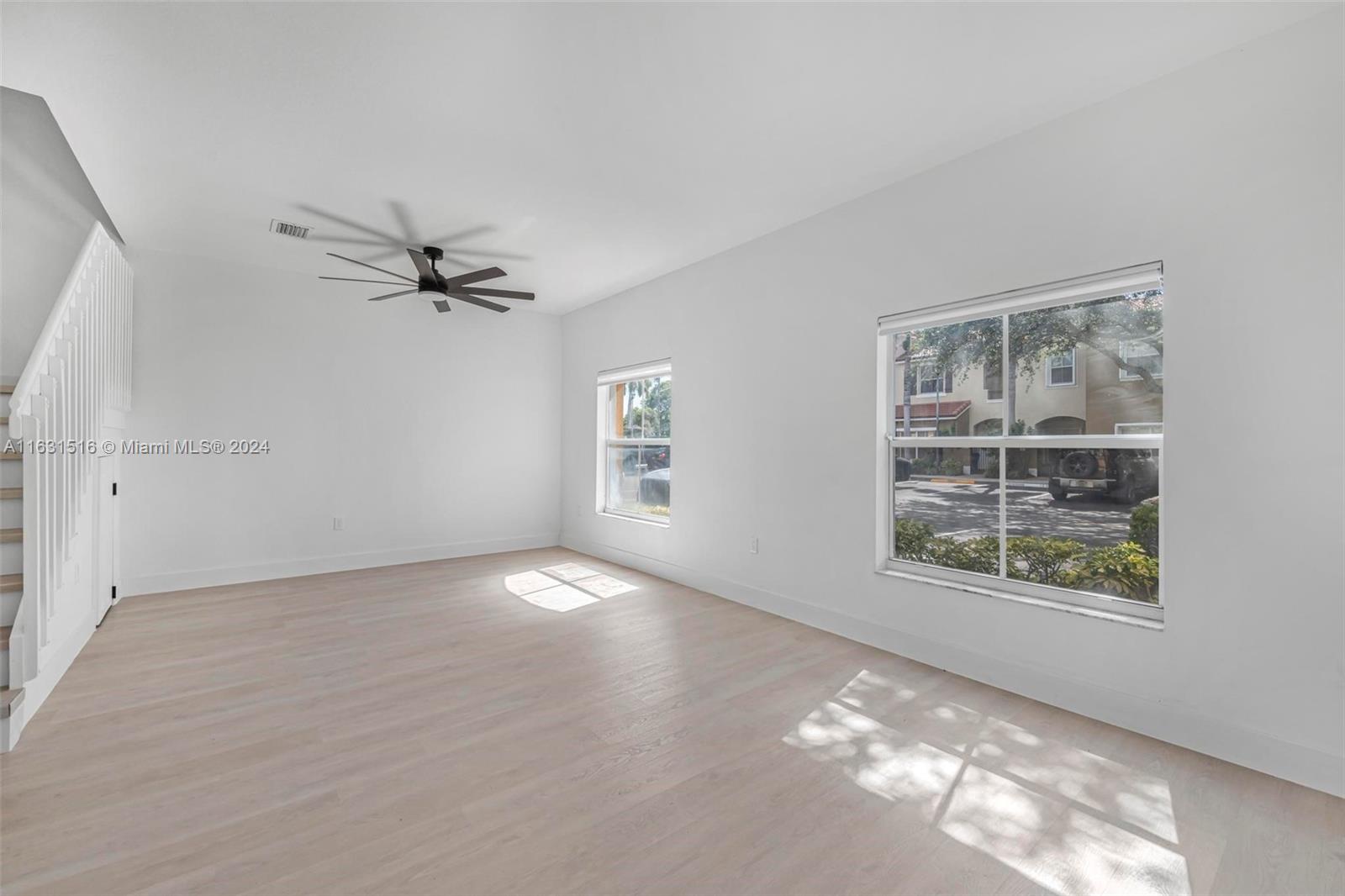 Vacant living room with pale wood floors, large windows, and a black ceiling fan.