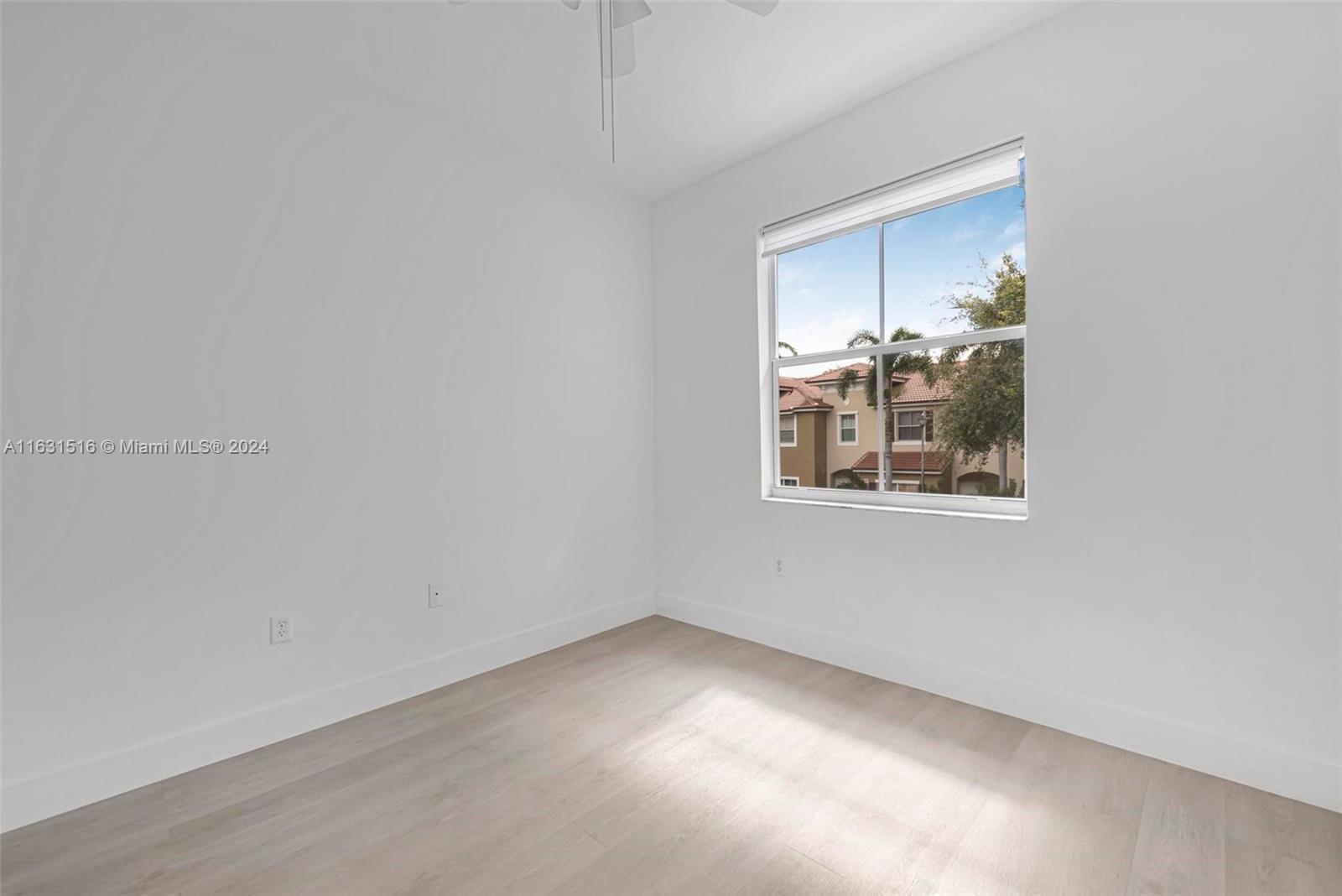 Empty bedroom with pale wood floors and a large window overlooking neighboring homes.