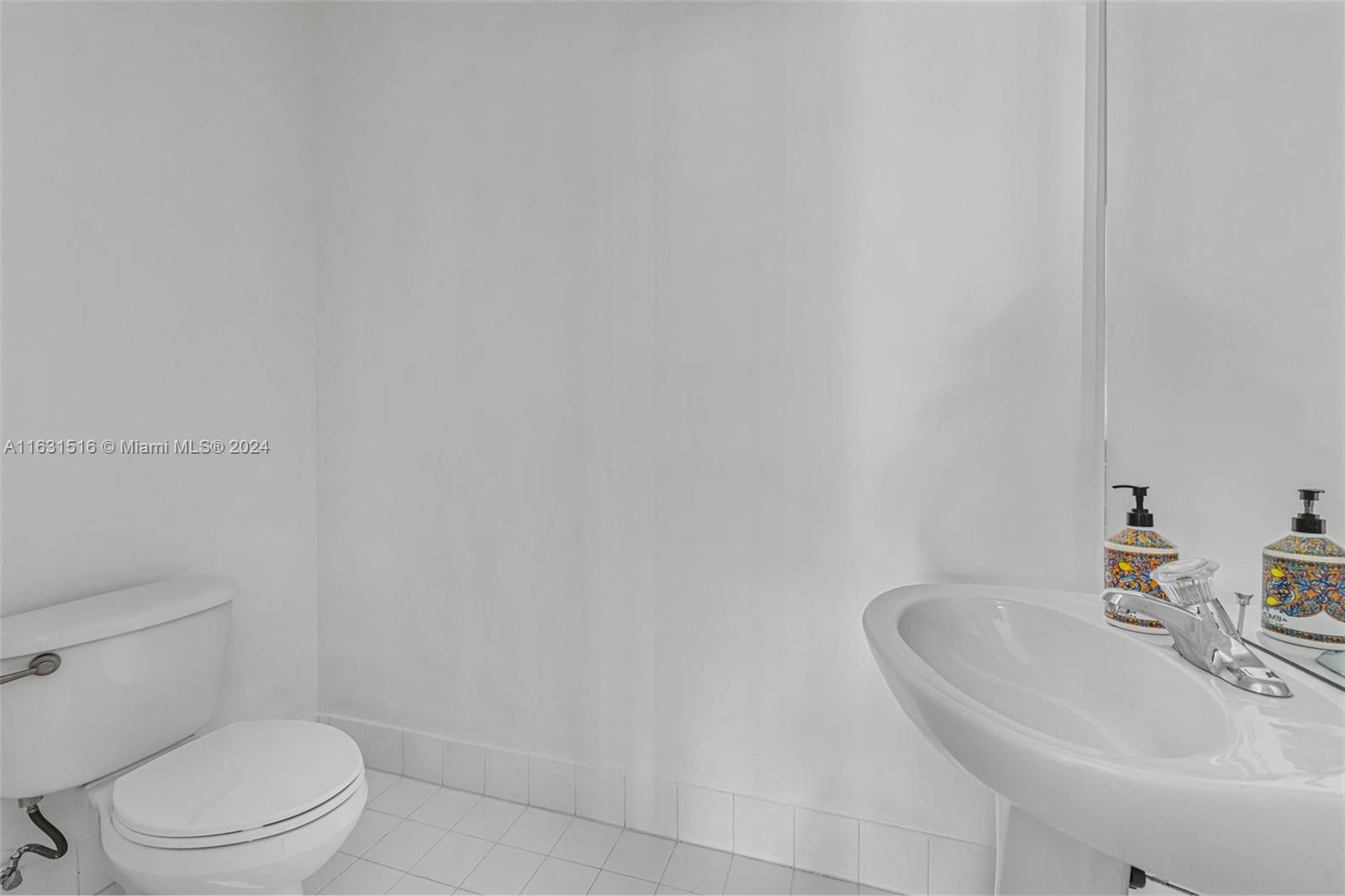 White bathroom with a toilet, oval sink, chrome faucet, and colorful soap dispensers.