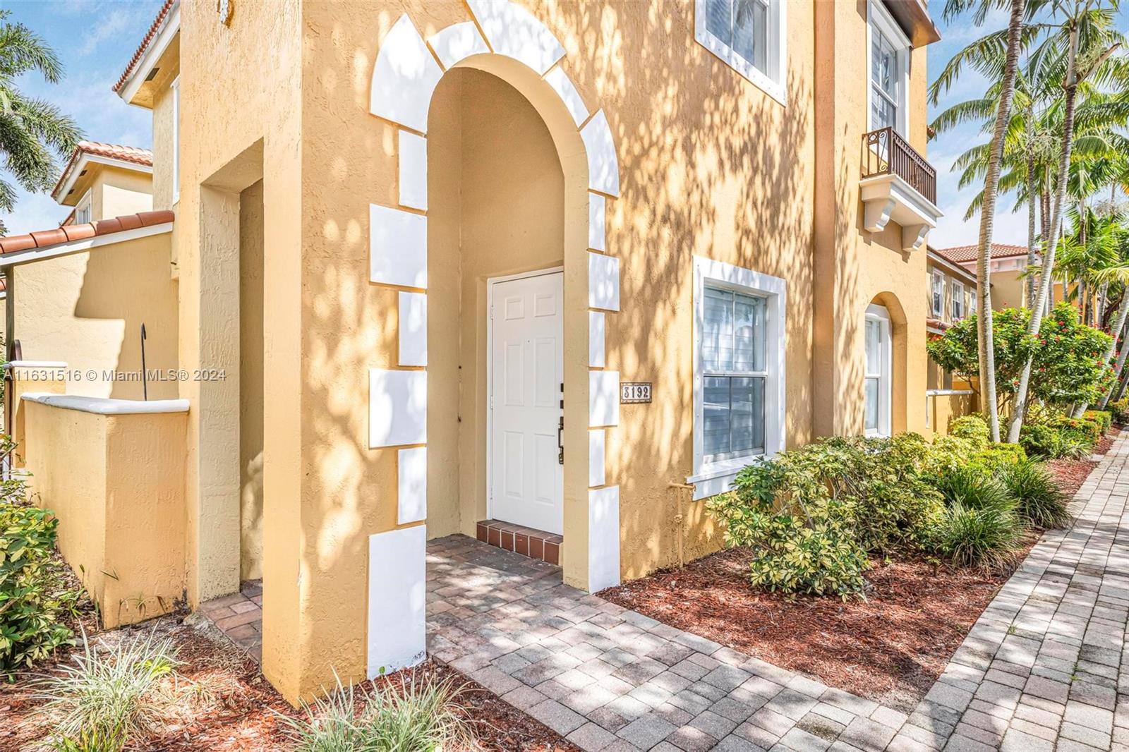 Yellow stucco residence with an arched entry, tropical landscaping, and a paved walkway.