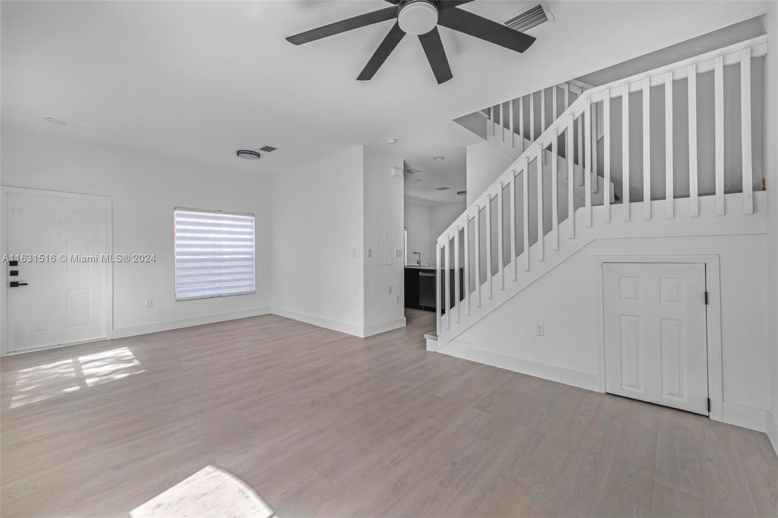 Vacant living room with light wood-look floors, a sweeping white staircase, and a dark ceiling fan.