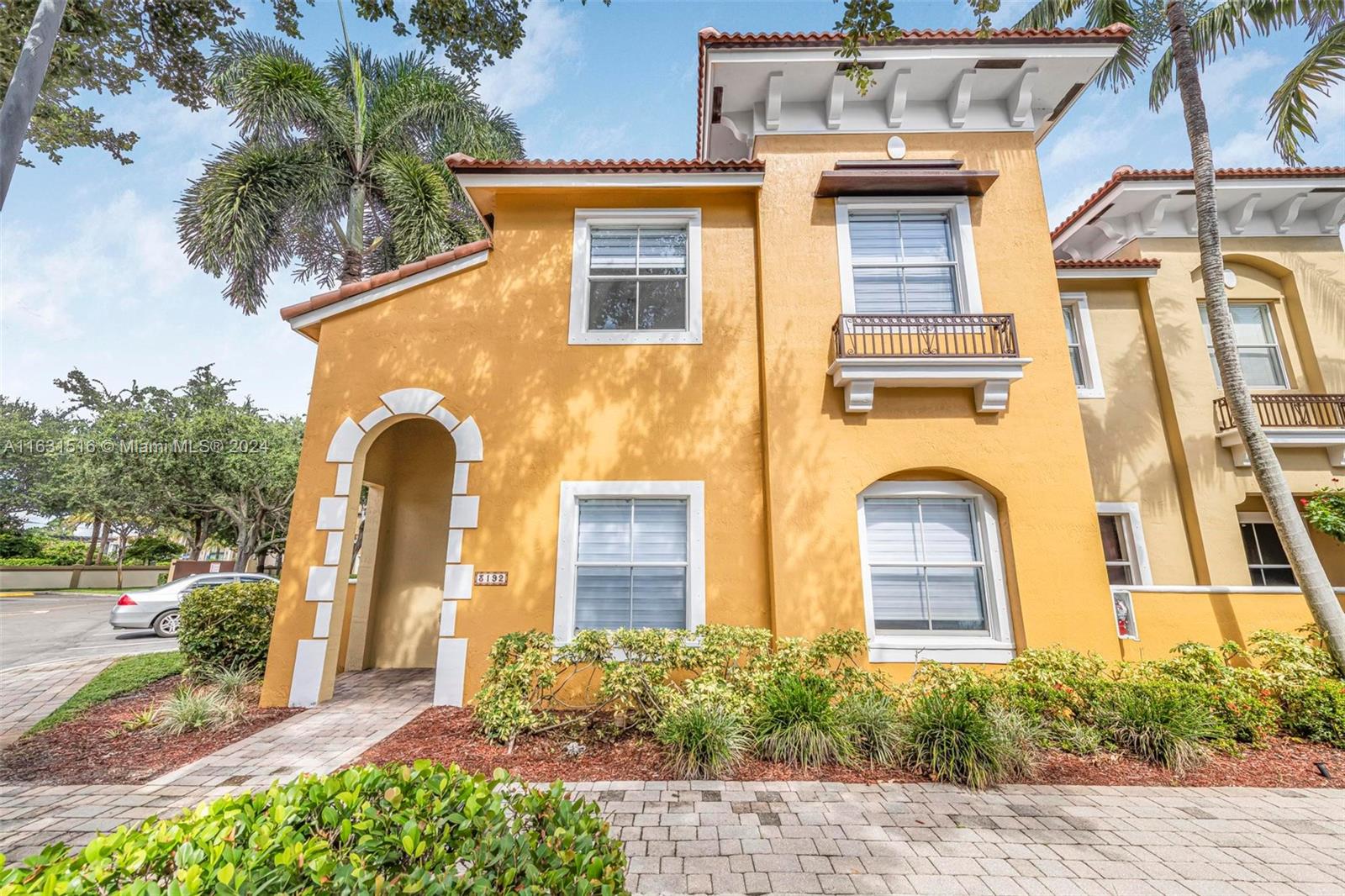 Yellow stucco townhouse with a tiled roof, small balcony, arched entrance, and tropical landscaping.