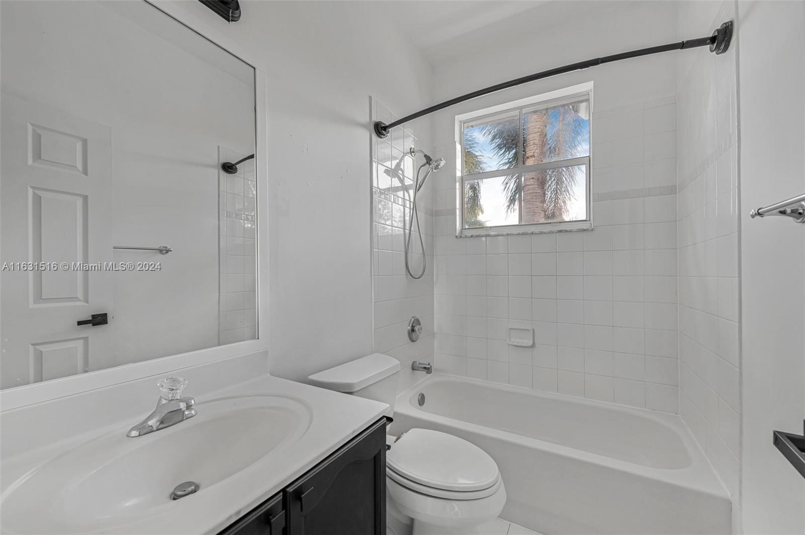 Bathroom with a white-tiled tub and shower, vanity sink, and window overlooking palm trees.