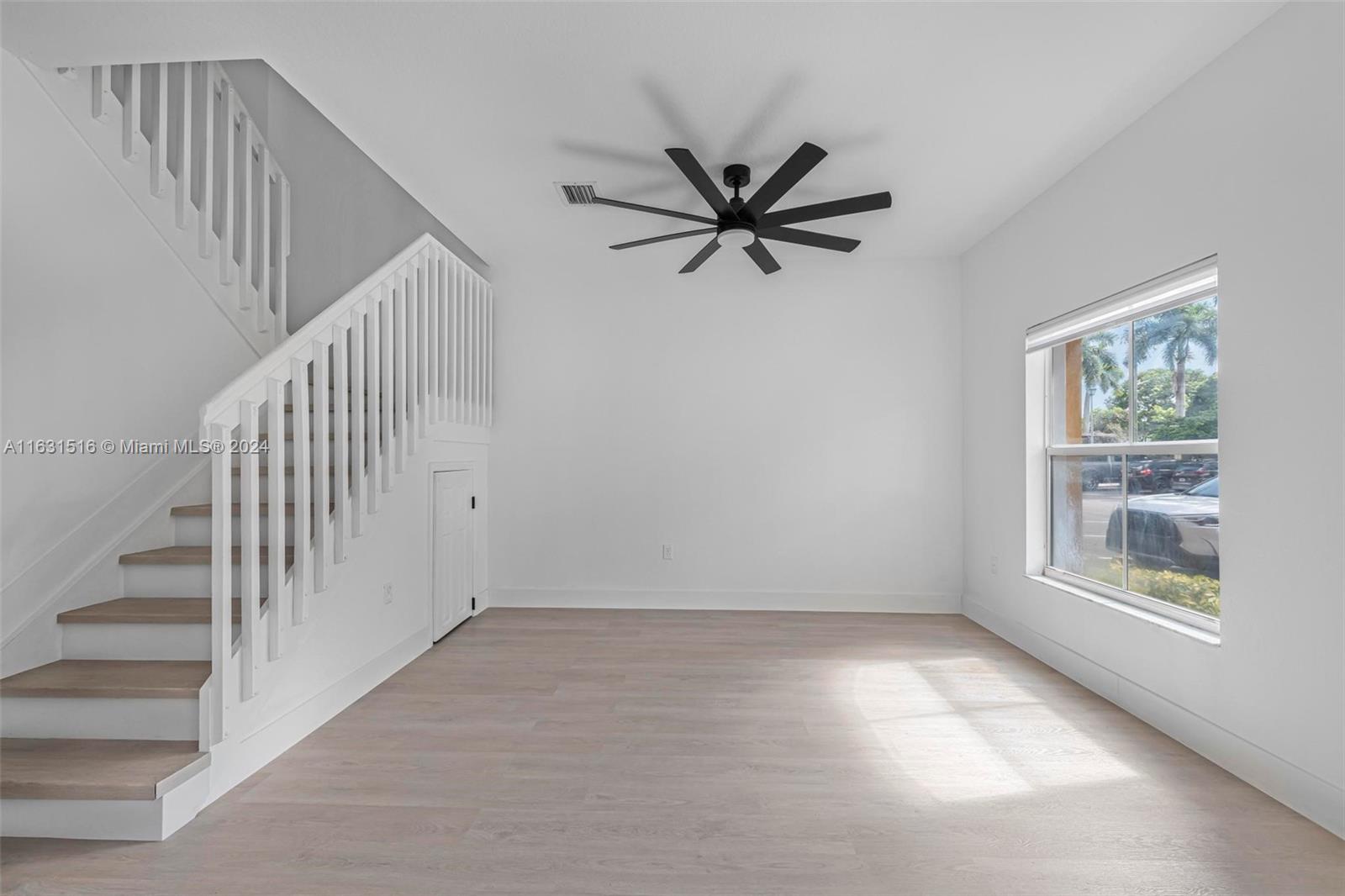 Vacant living area with a white-railed staircase, pale wood floors, and a large window.