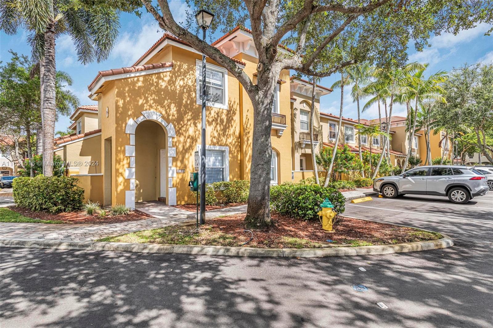 Yellow stucco condominium building with an arched entry, red tile roof, and palm-lined parking area.