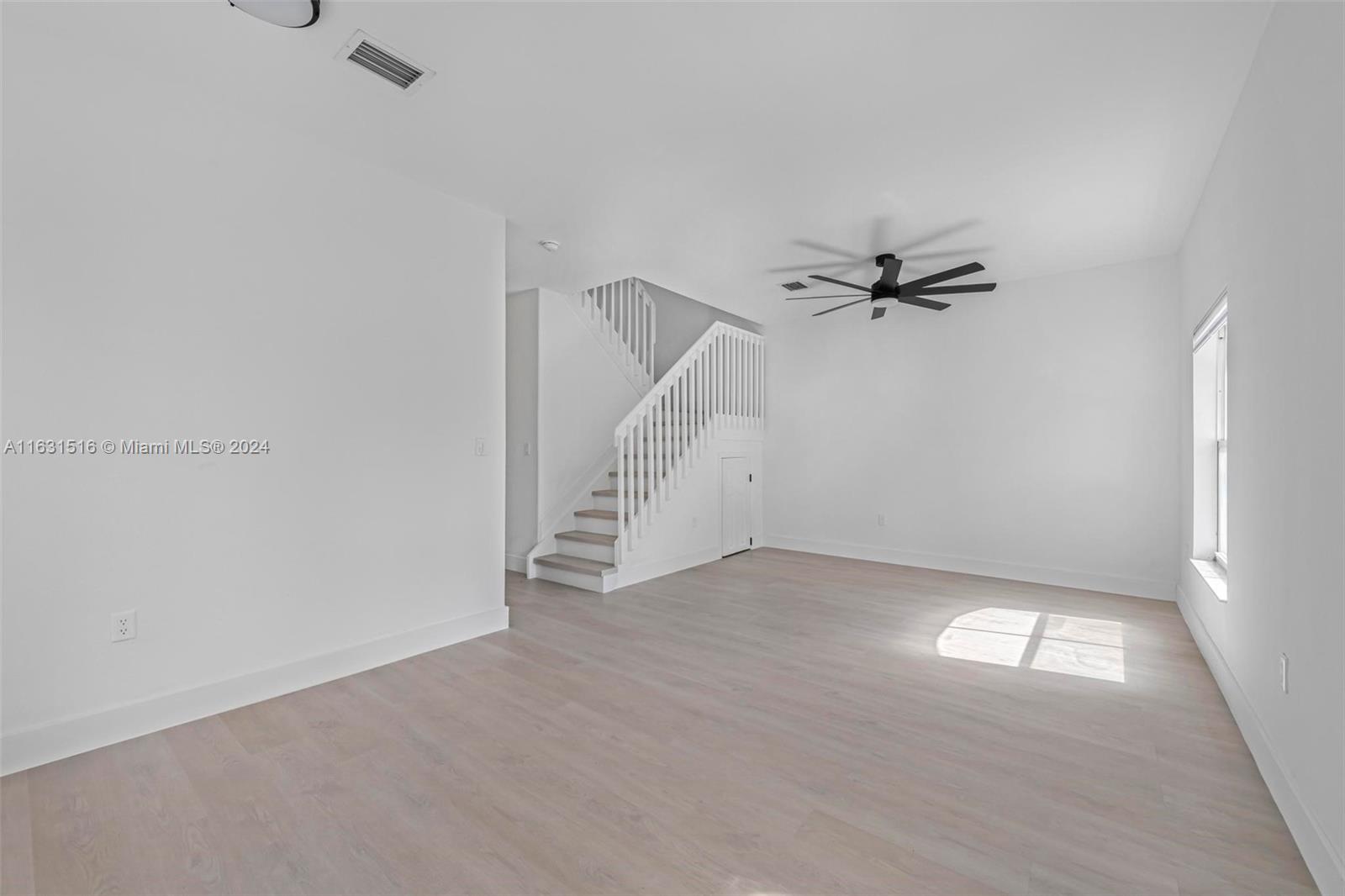 Empty living room with pale flooring, a black ceiling fan, and a white-railed staircase.