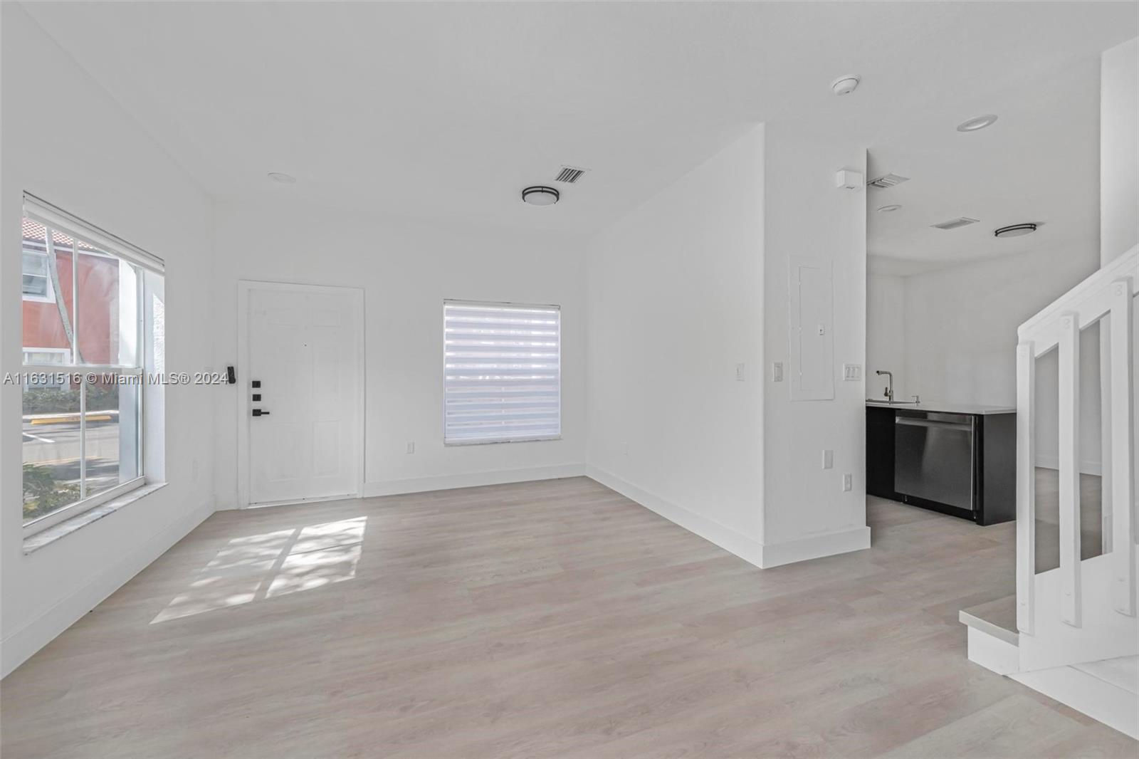 Vacant living area with pale flooring, a large window, compact kitchen, and white staircase railing.
