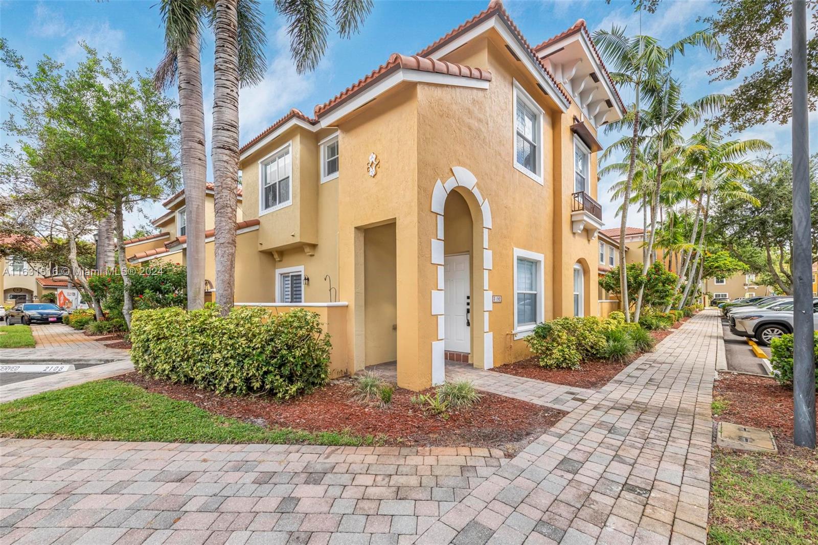 Yellow two-story townhouse with an arched entry, tile roof, and palm-lined paved walkway.