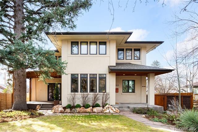 Modern beige two-story home with broad rooflines, a covered entry, and a large evergreen in the front yard.