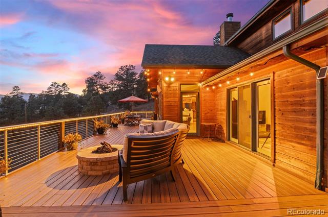 Wooden home and deck with a fire pit and lounge seating overlooking trees at dusk.