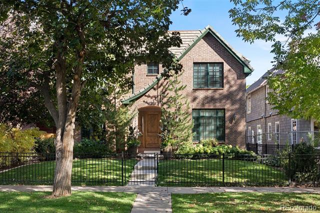 Two-story brick house with a steep gabled roof, mature trees, landscaped lawn, and black metal fence.