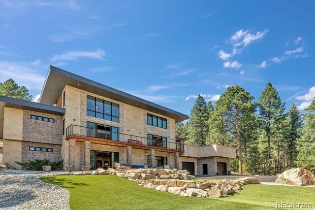 Modern stone-and-wood home with a balcony, landscaped lawn, and pine trees.