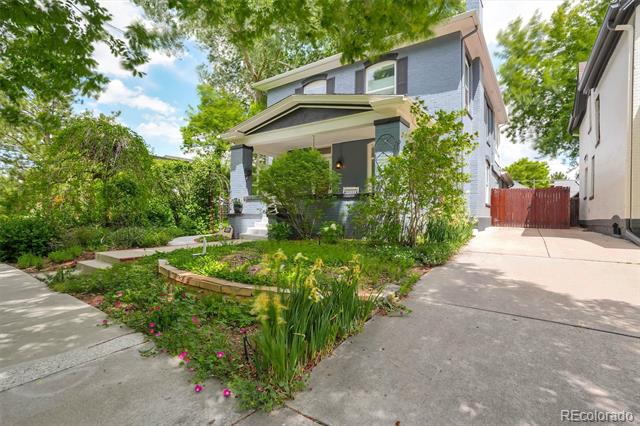 Two-story blue-gray house with a covered porch, flower garden, and concrete driveway.