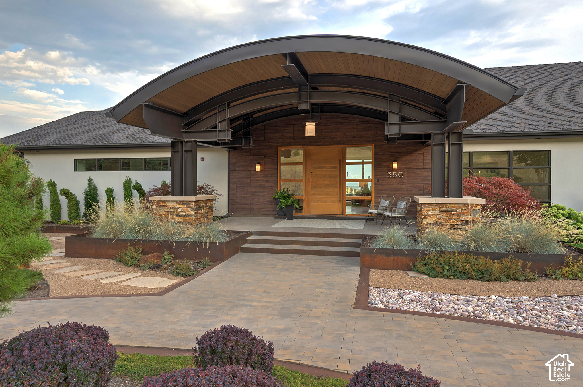 Modern home entrance with a curved metal canopy, wood-paneled facade, and landscaped paver walkway.