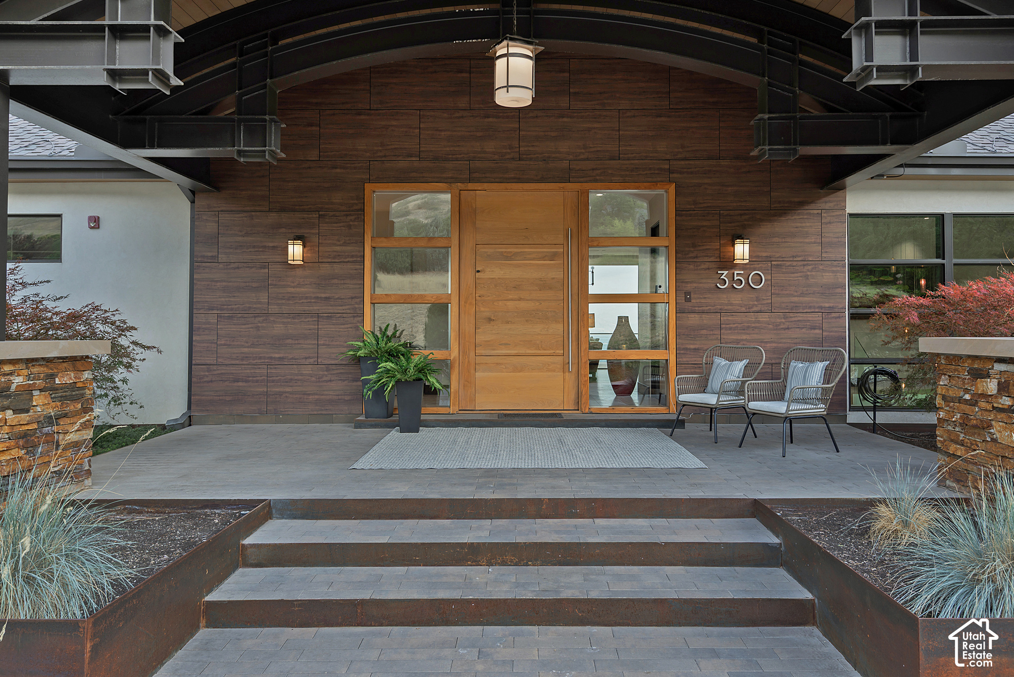 Modern home entrance with a tall wood door, dark wood facade, covered porch, and broad paved steps.