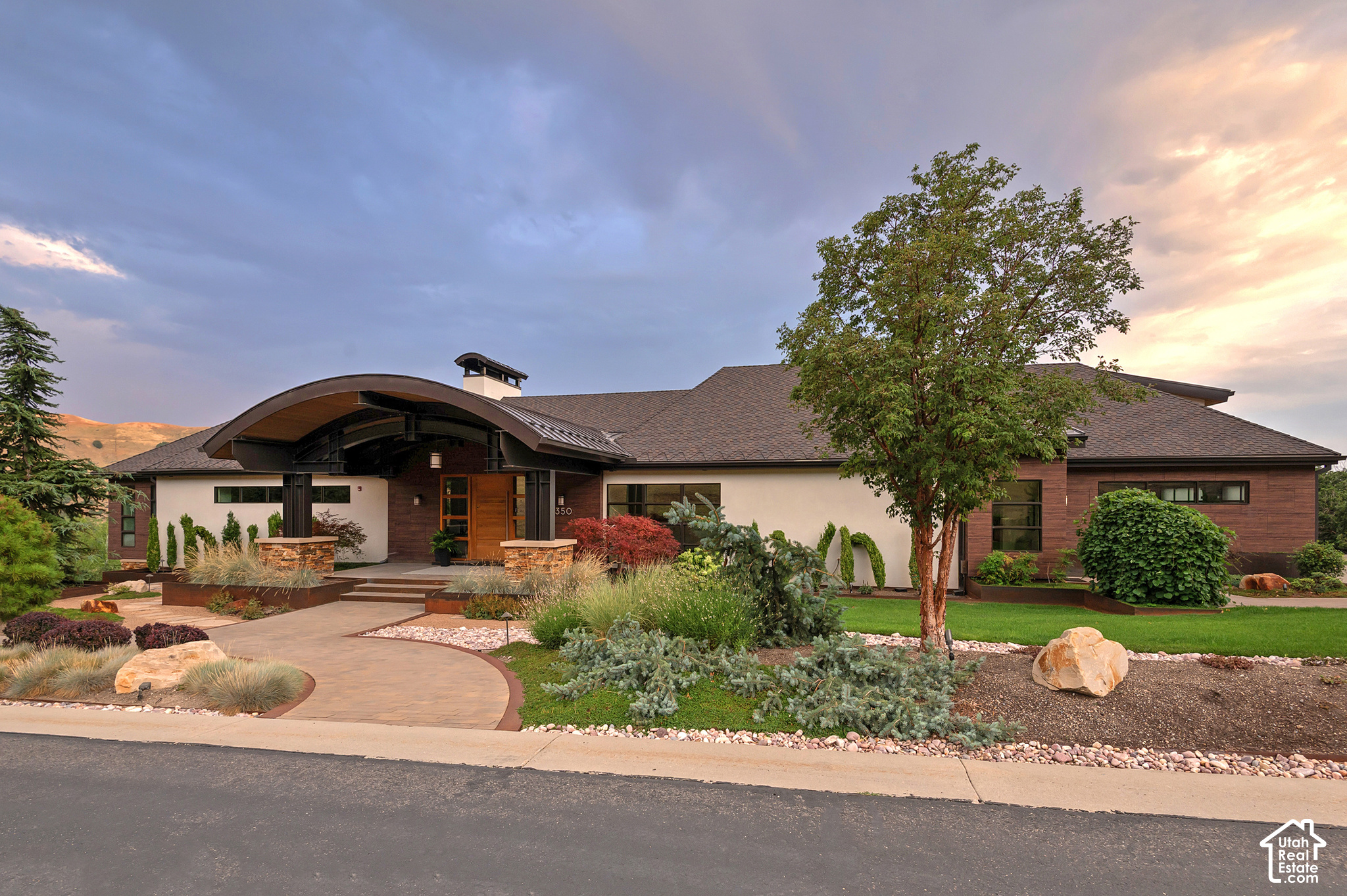 Modern home with a curved covered entry and landscaped front garden at sunset.