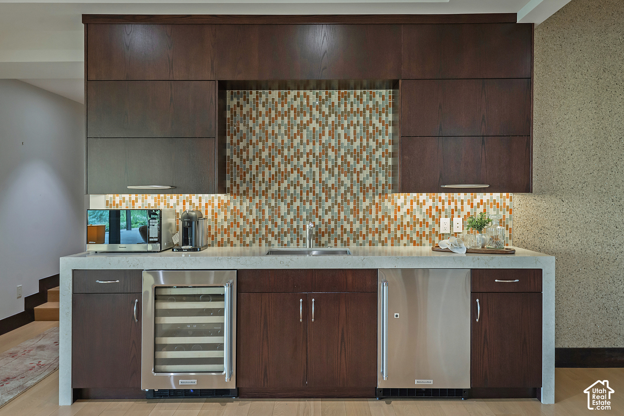 Modern kitchen wet bar with dark wood cabinets, colorful tile backsplash, and stainless-steel appliances.