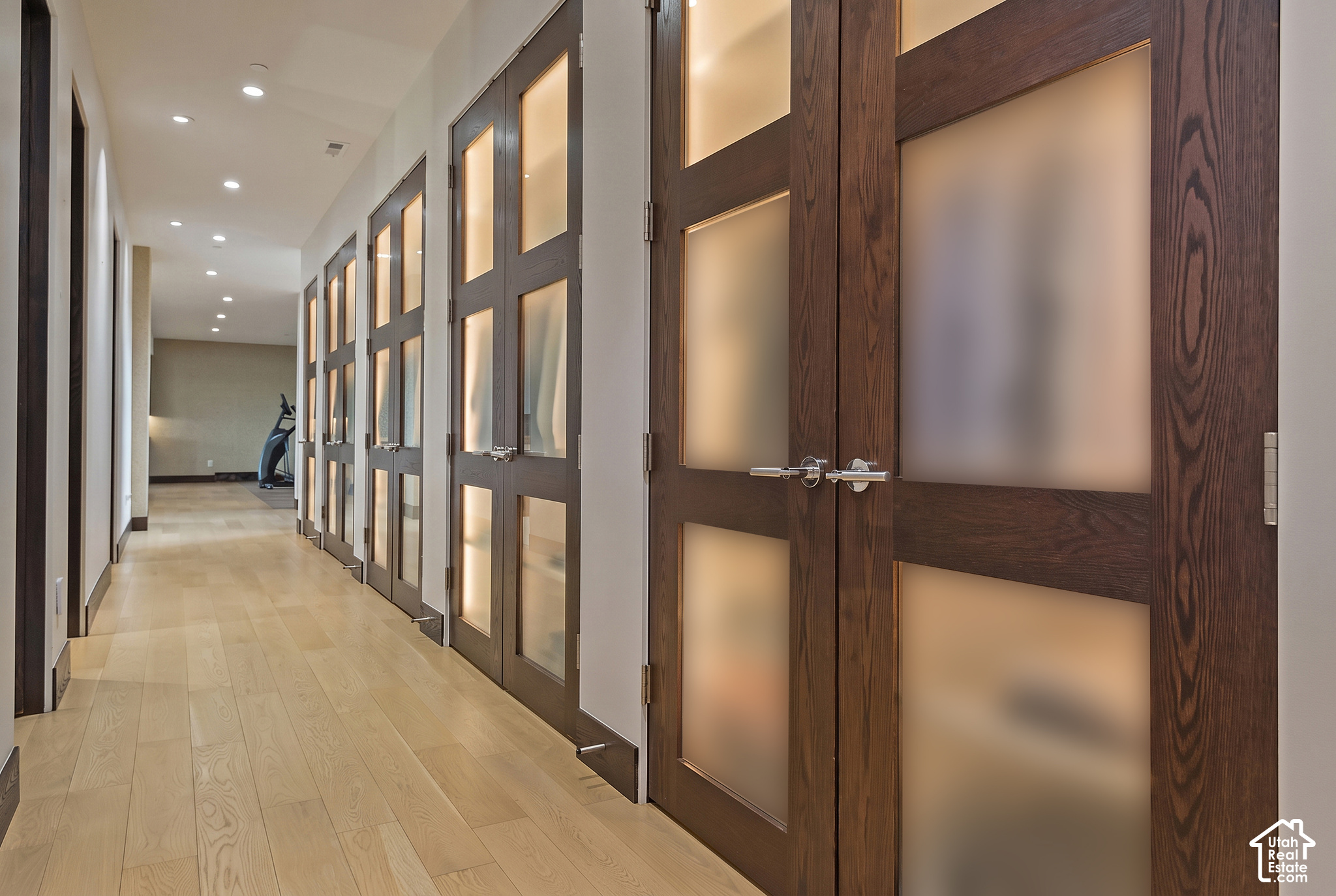 Interior hallway with pale wood floors, dark wood doors, and exercise equipment at the far end.