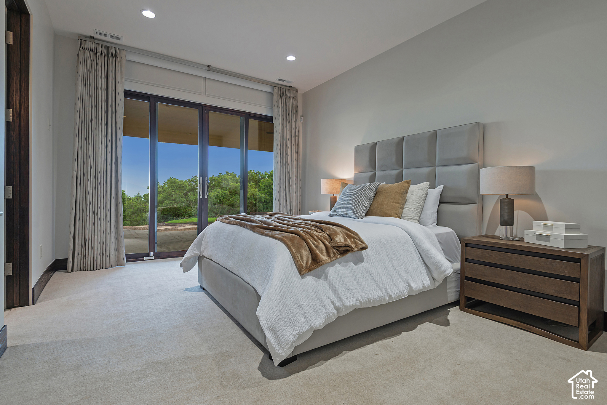 Staged bedroom with a large upholstered bed beside sliding glass doors overlooking leafy trees.