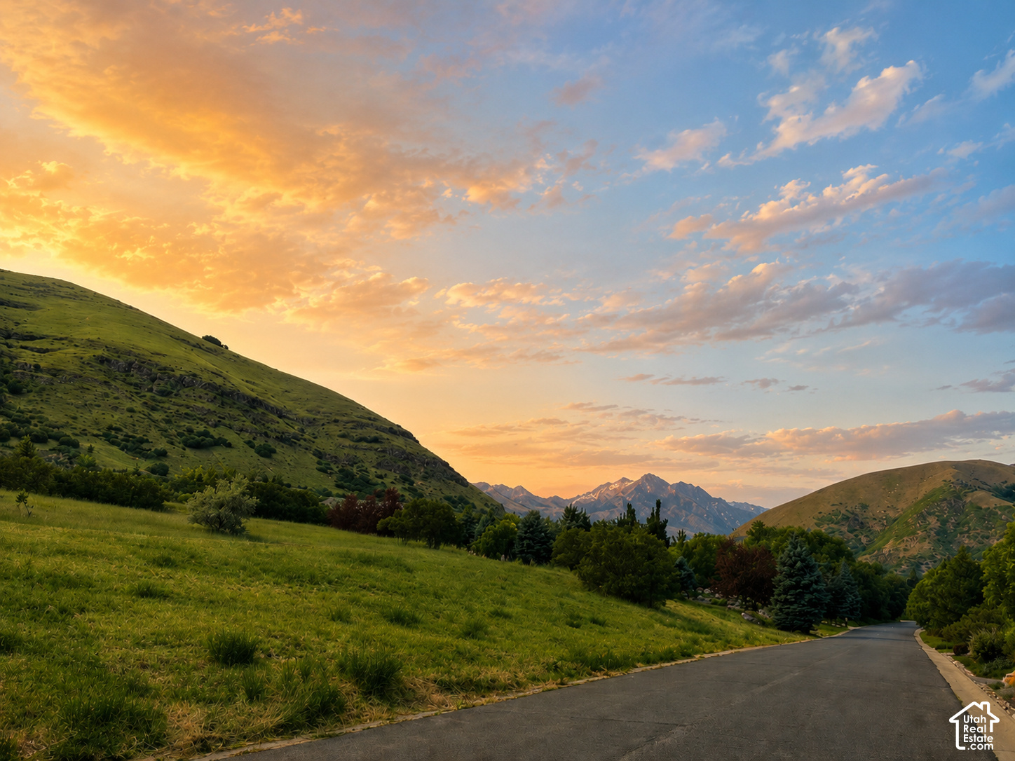 A winding road between grassy foothills with distant mountains beneath a sunset sky.