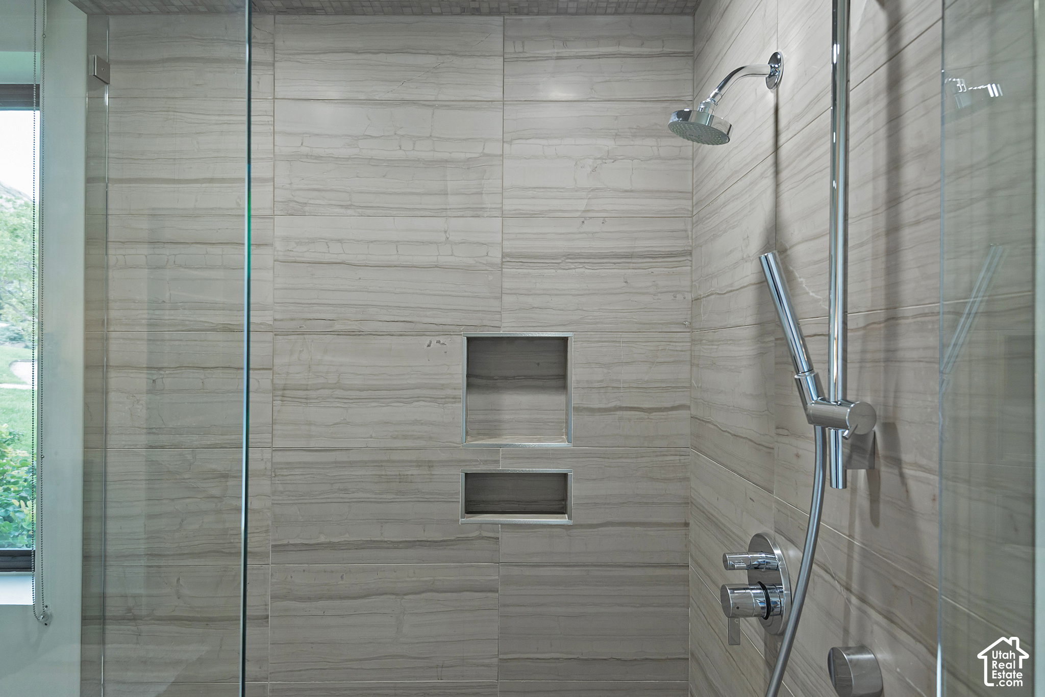 Walk-in shower with gray stone tile, two recessed niches, and chrome fixtures beside a window.