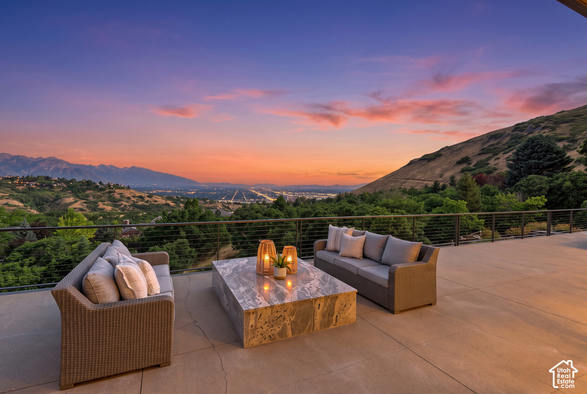 Furnished patio with wicker seating overlooks Salt Lake City and mountains beneath a colorful sunset.