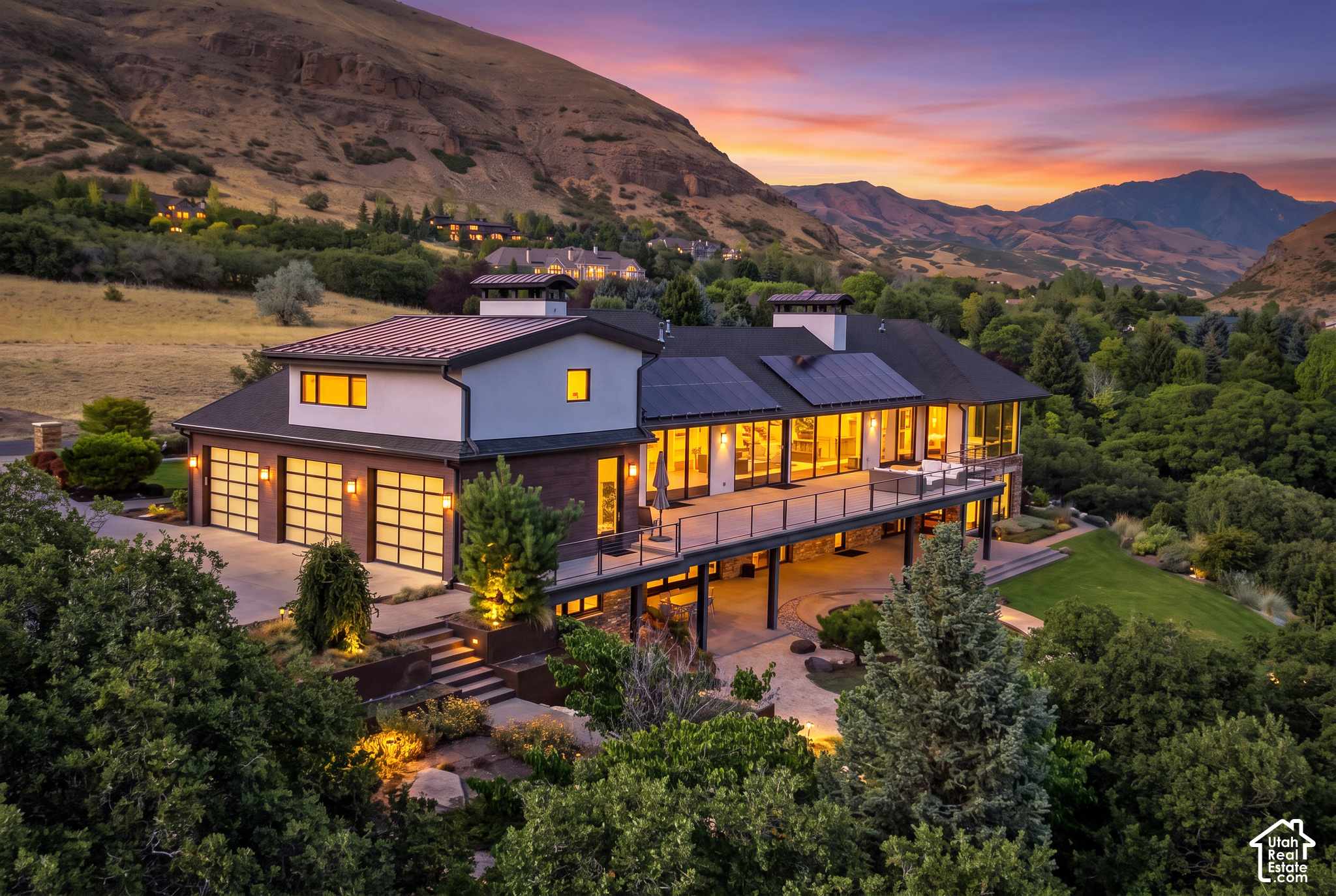 Modern home with solar panels and a glass-fronted terrace, set against mountains at dusk.