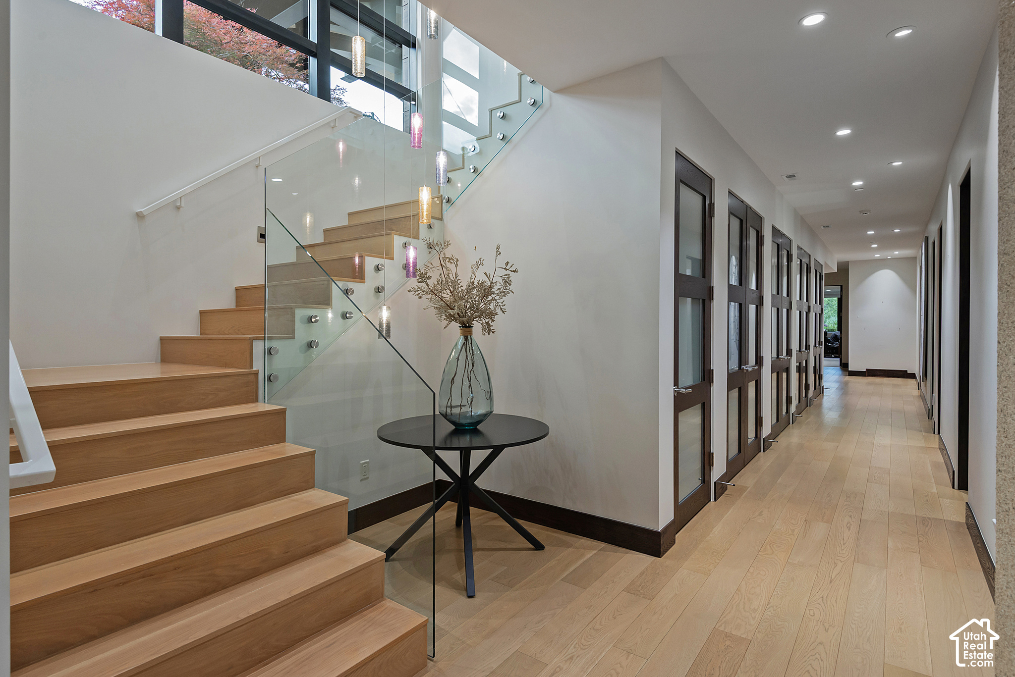 Modern entry hall with a wood staircase, glass railing, and long corridor of dark-framed doors.
