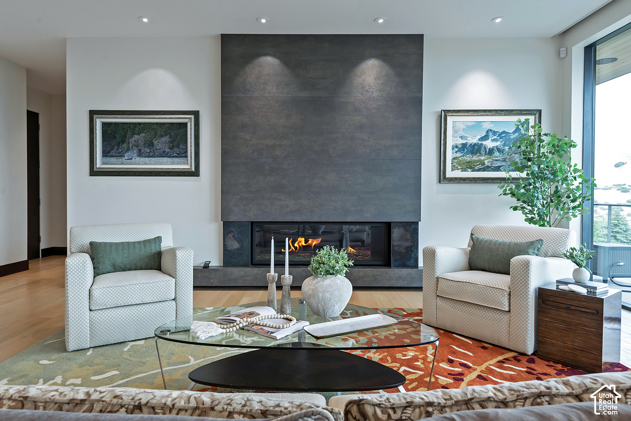 Living room with a tall stone fireplace, lit fire, patterned armchairs, and large windows.