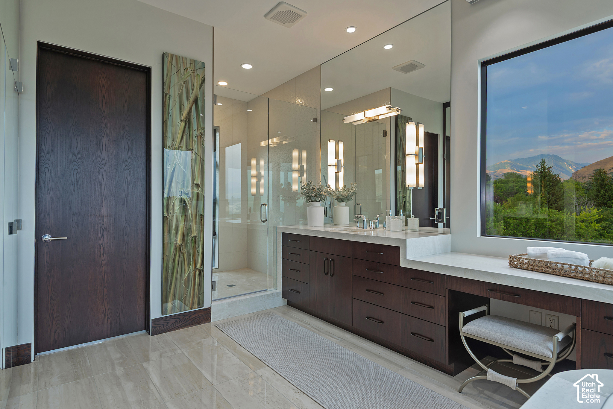 Modern bathroom with a glass shower, dark double vanity, and mountain views through a large window.