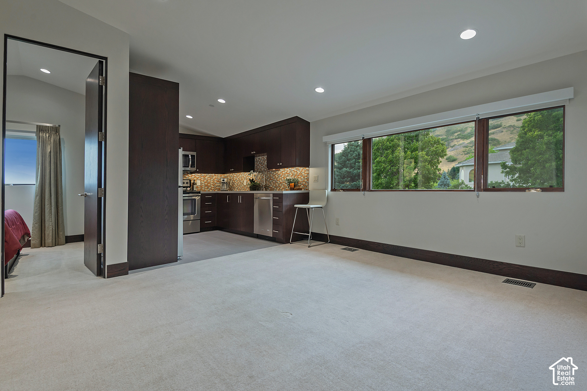 Open carpeted living area beside a dark-wood kitchen with a long window overlooking trees.