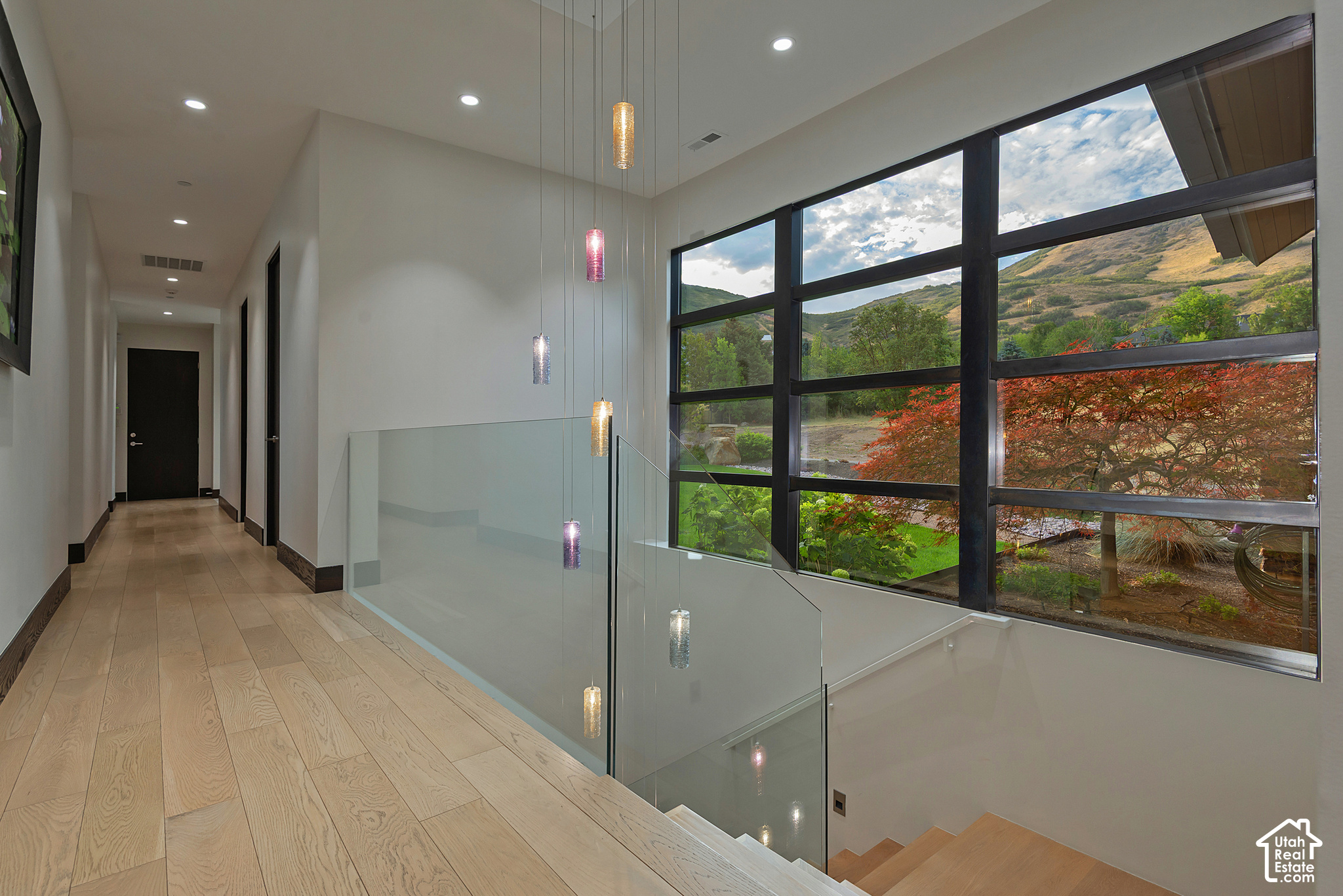 Modern hallway and stair landing with glass railing, pendant lights, and large windows overlooking green hills.