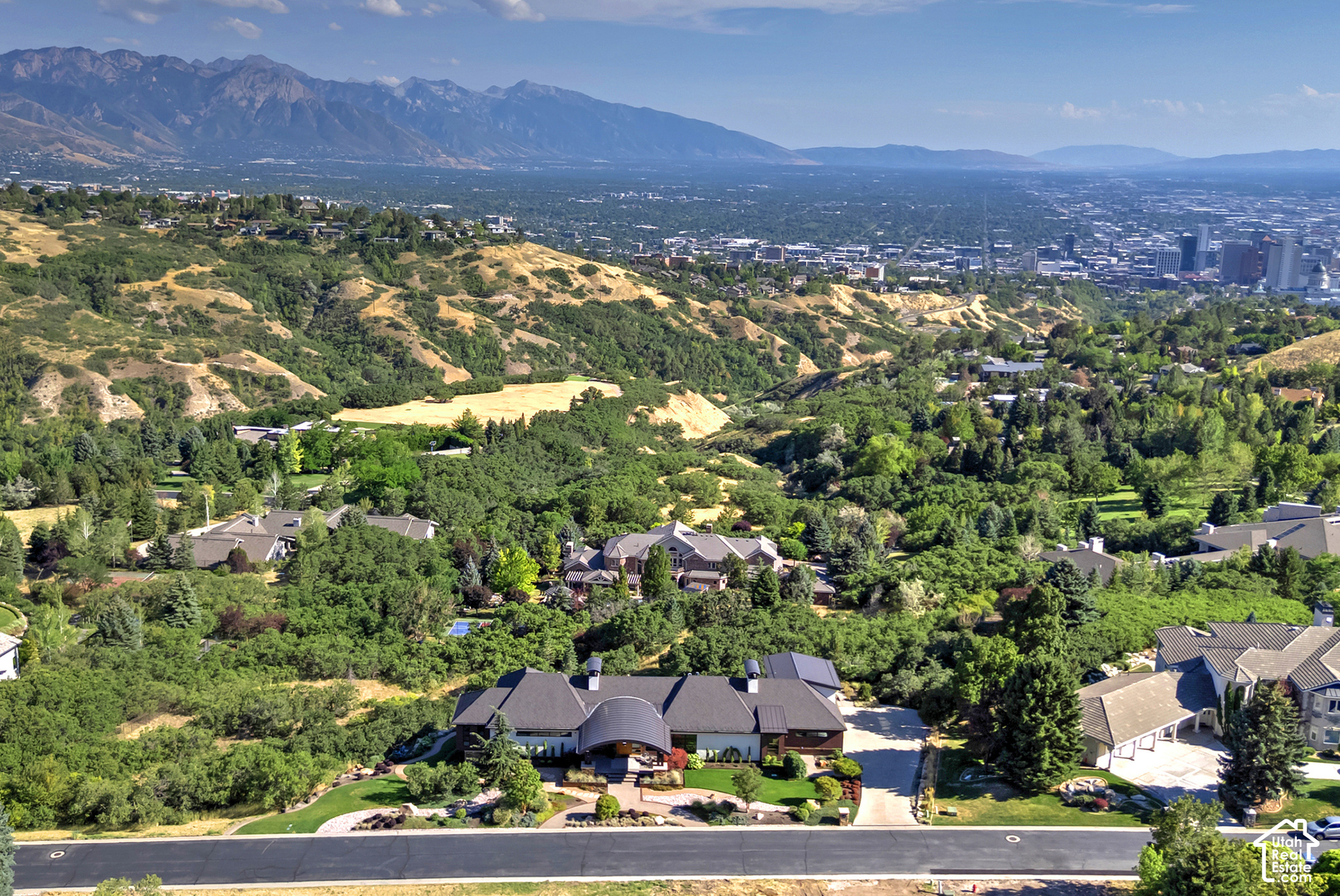 Aerial view of a large home, wooded hills, Salt Lake City skyline, and distant mountains.