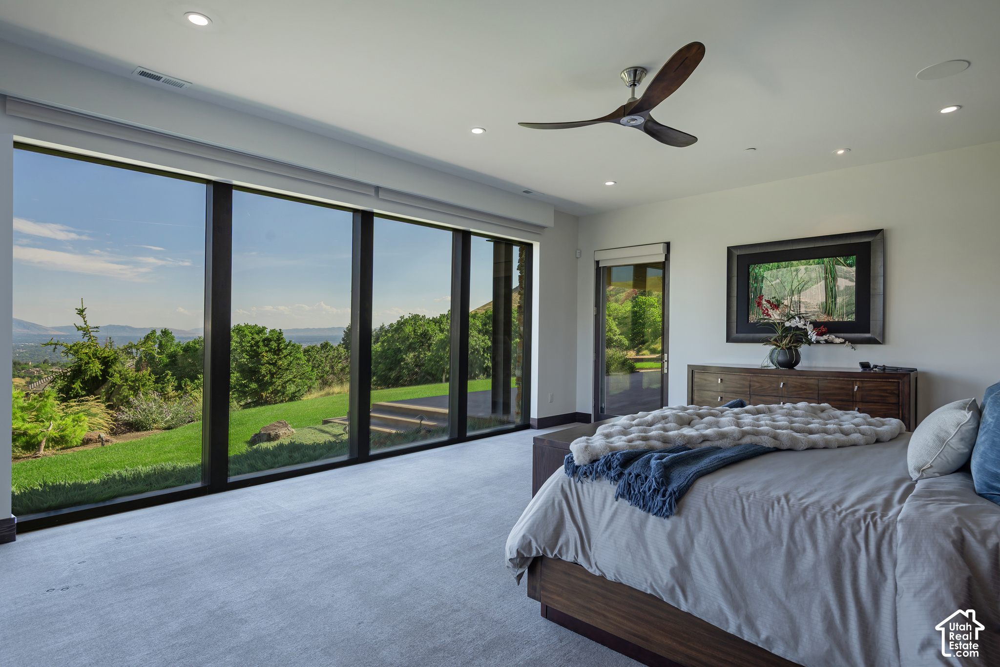 Modern bedroom with floor-to-ceiling glass doors overlooking a landscaped yard and distant hills.