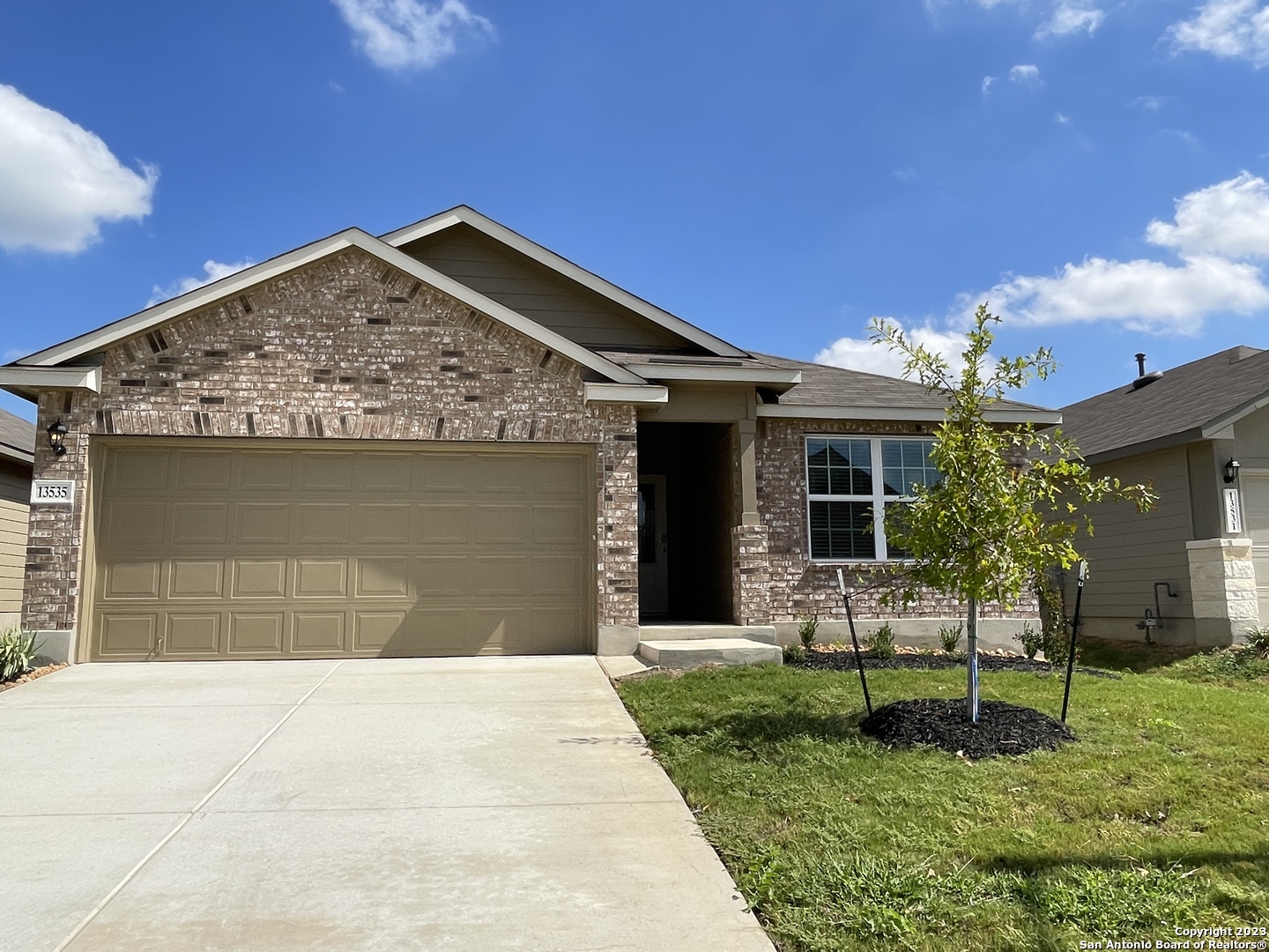 Single-story brick house with a two-car garage, wide driveway, and young tree in the front lawn.