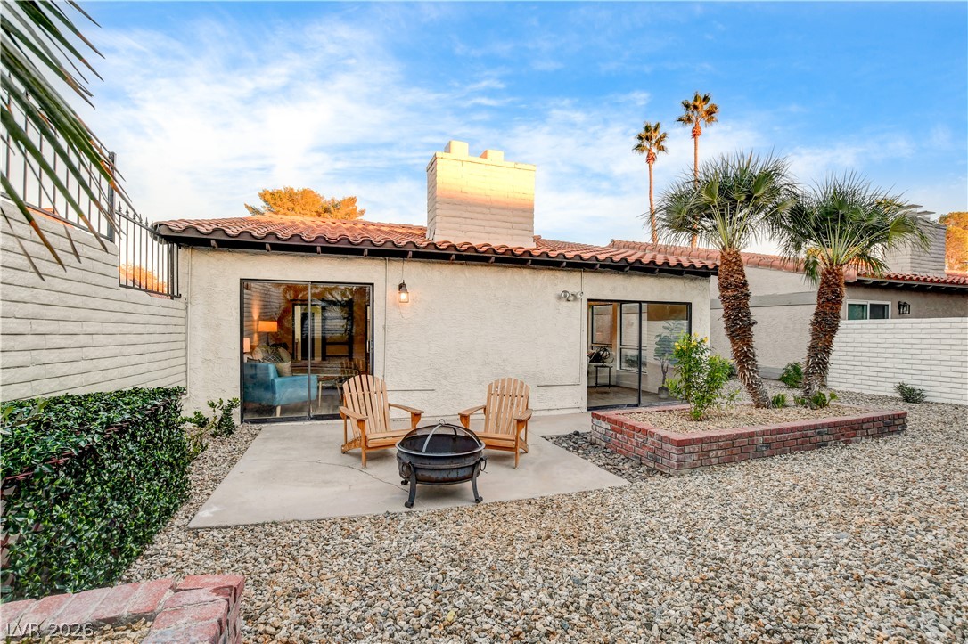 Home’s rear patio with two wooden chairs, a fire pit, gravel landscaping, and palm trees.
