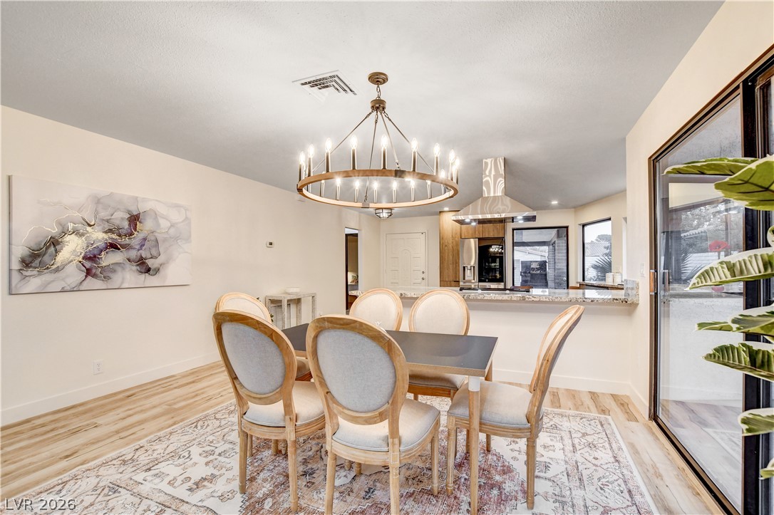 Dining area with a circular chandelier, upholstered chairs, abstract wall art, and an open kitchen behind it.