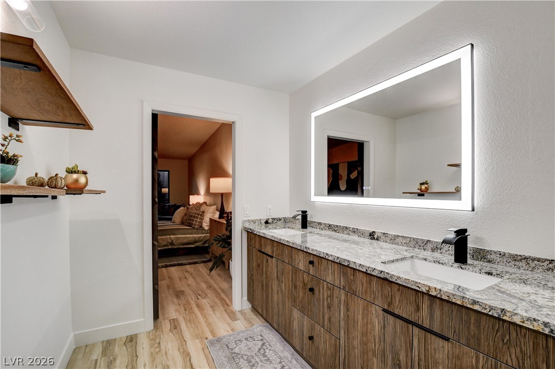 Bathroom with a long double vanity, stone countertop, and illuminated mirror beside an open bedroom doorway.