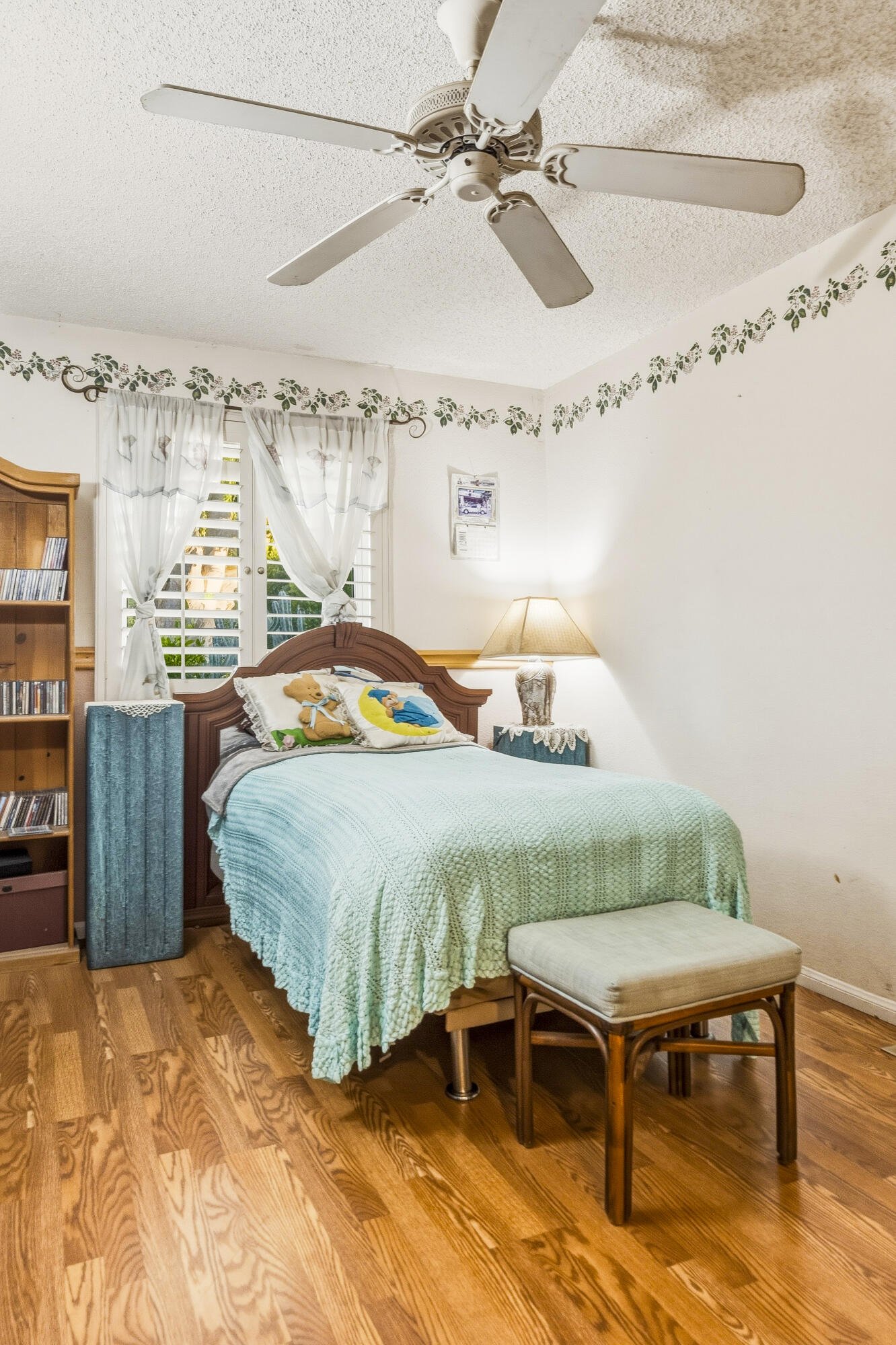 Bedroom with a turquoise quilt, hardwood floors, plantation shutters, and a ceiling fan.
