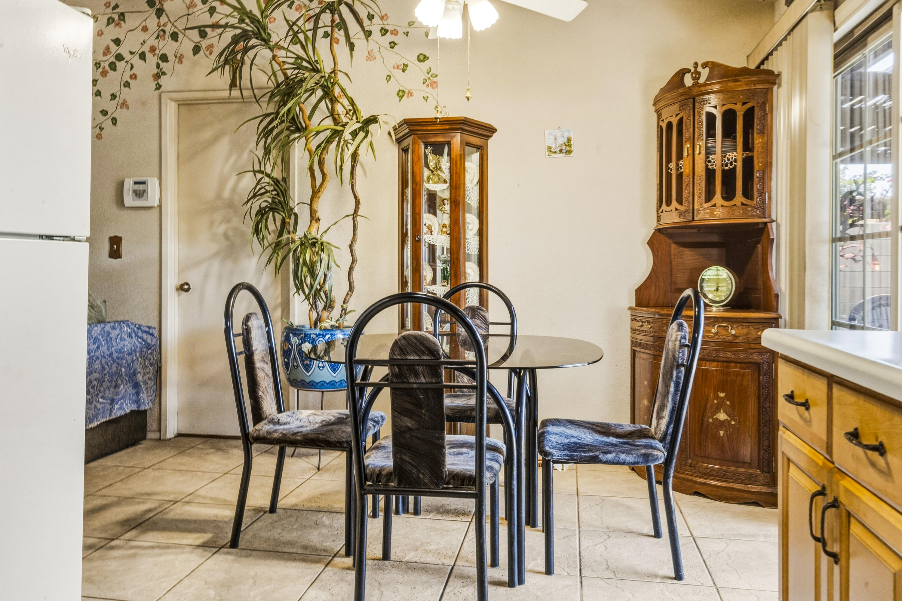 Dining area with a glass-top table, tall plant, and ornate wooden hutch beside the kitchen.