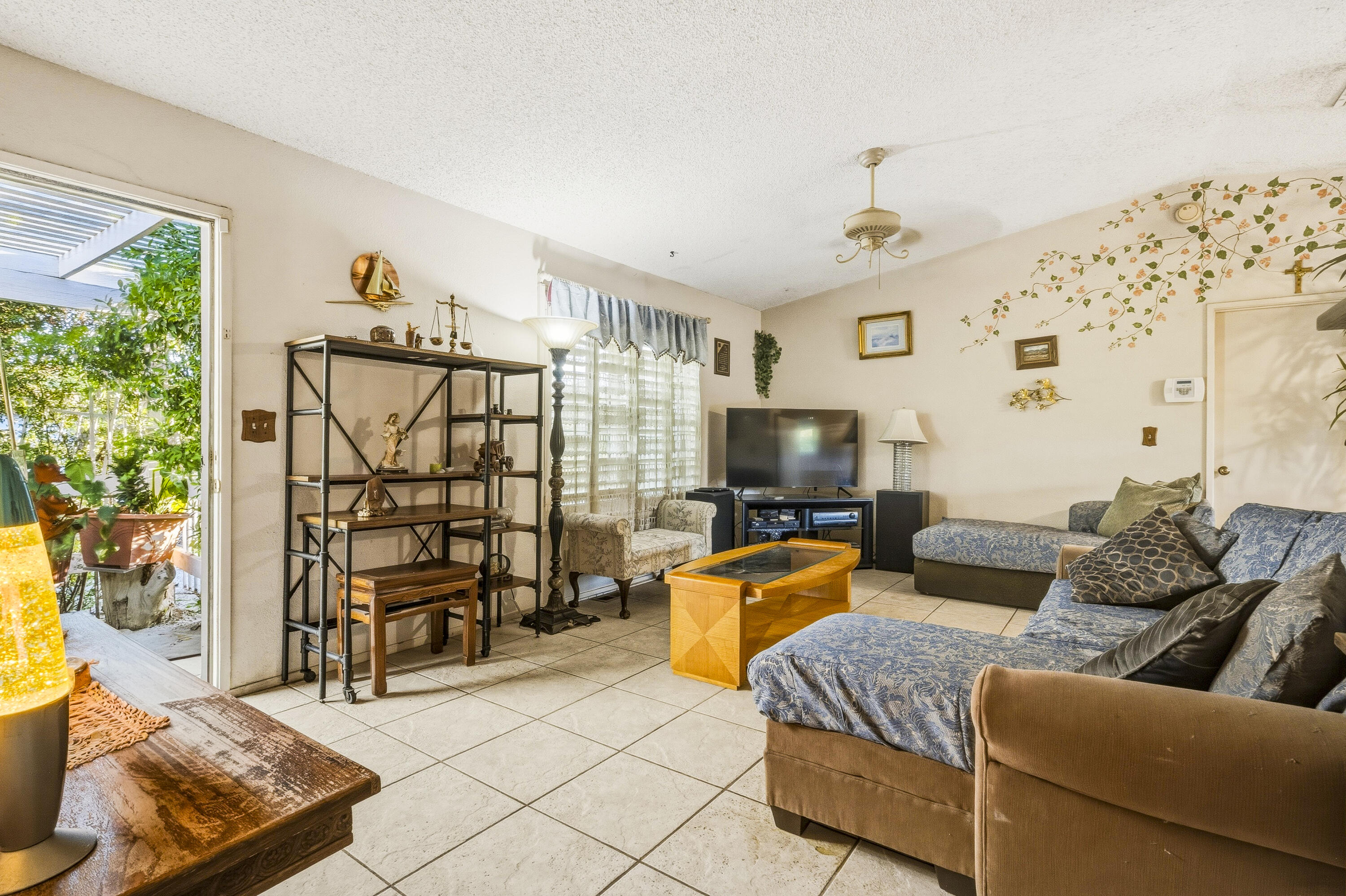 Living room with blue-patterned sofas, tiled floors, and sliding doors opening onto a leafy patio.