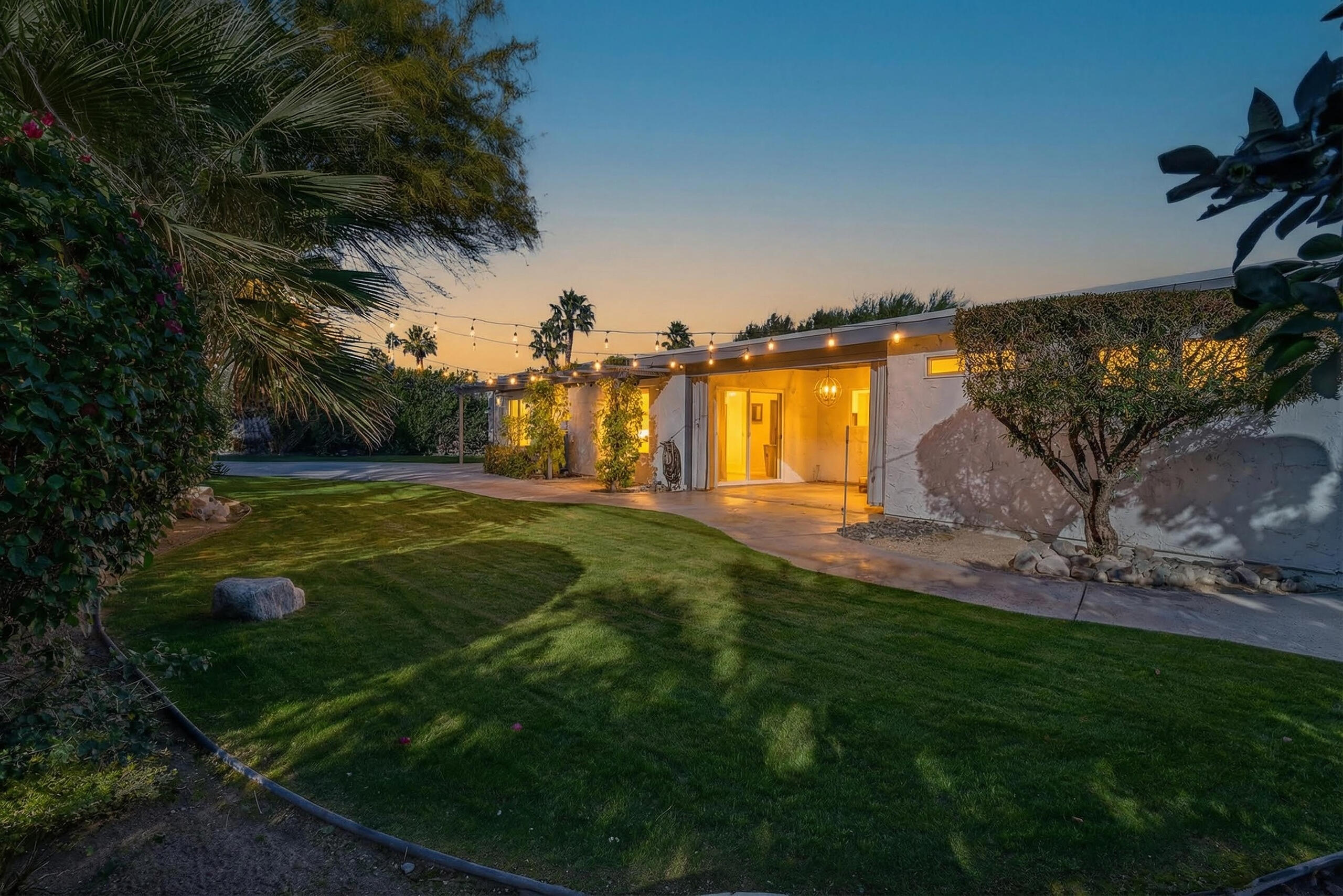 Single-story home with an illuminated patio and string lights beside a spacious lawn at dusk.