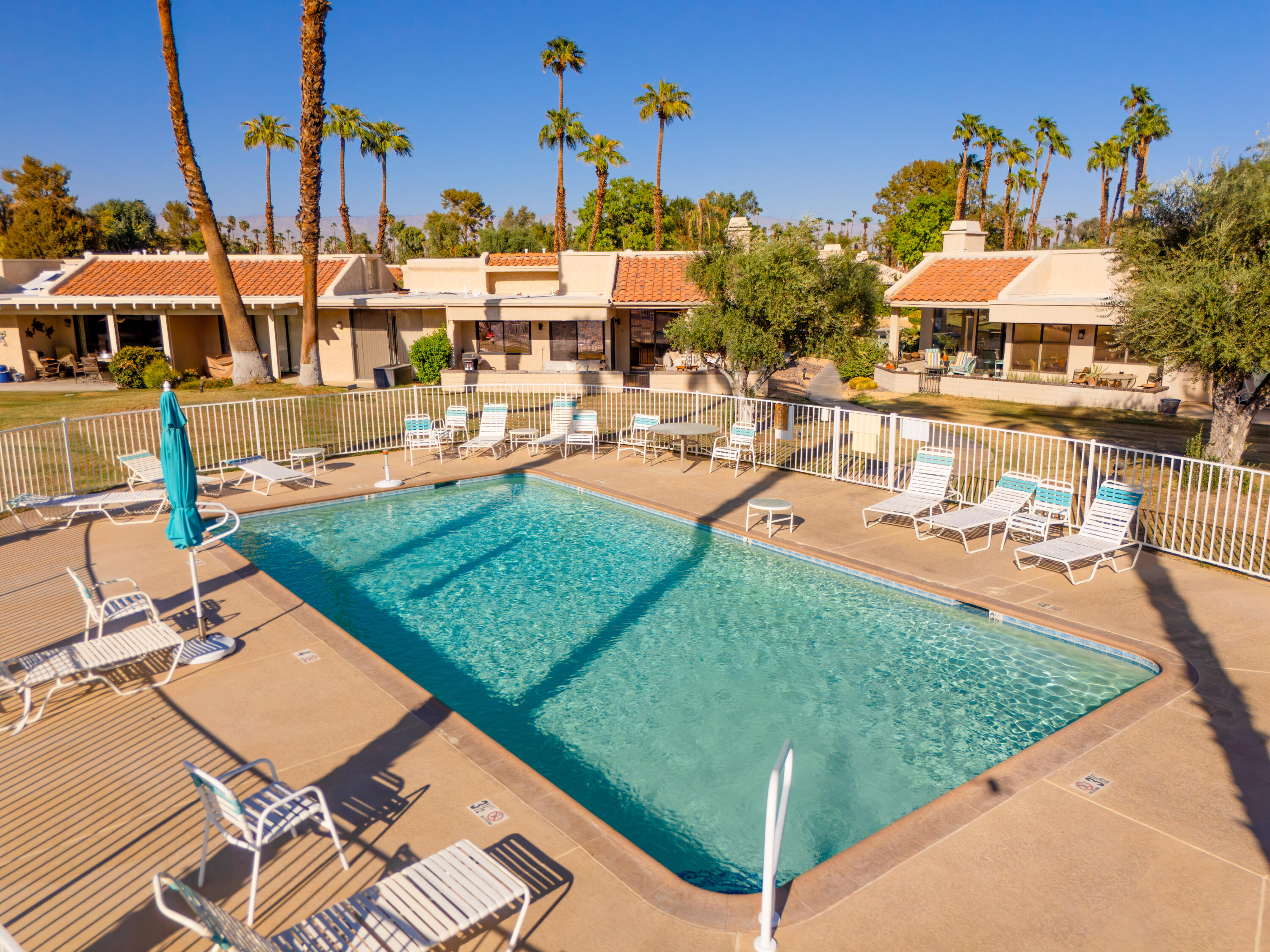 Outdoor swimming pool with lounge chairs, a turquoise umbrella and tile-roofed homes behind it.