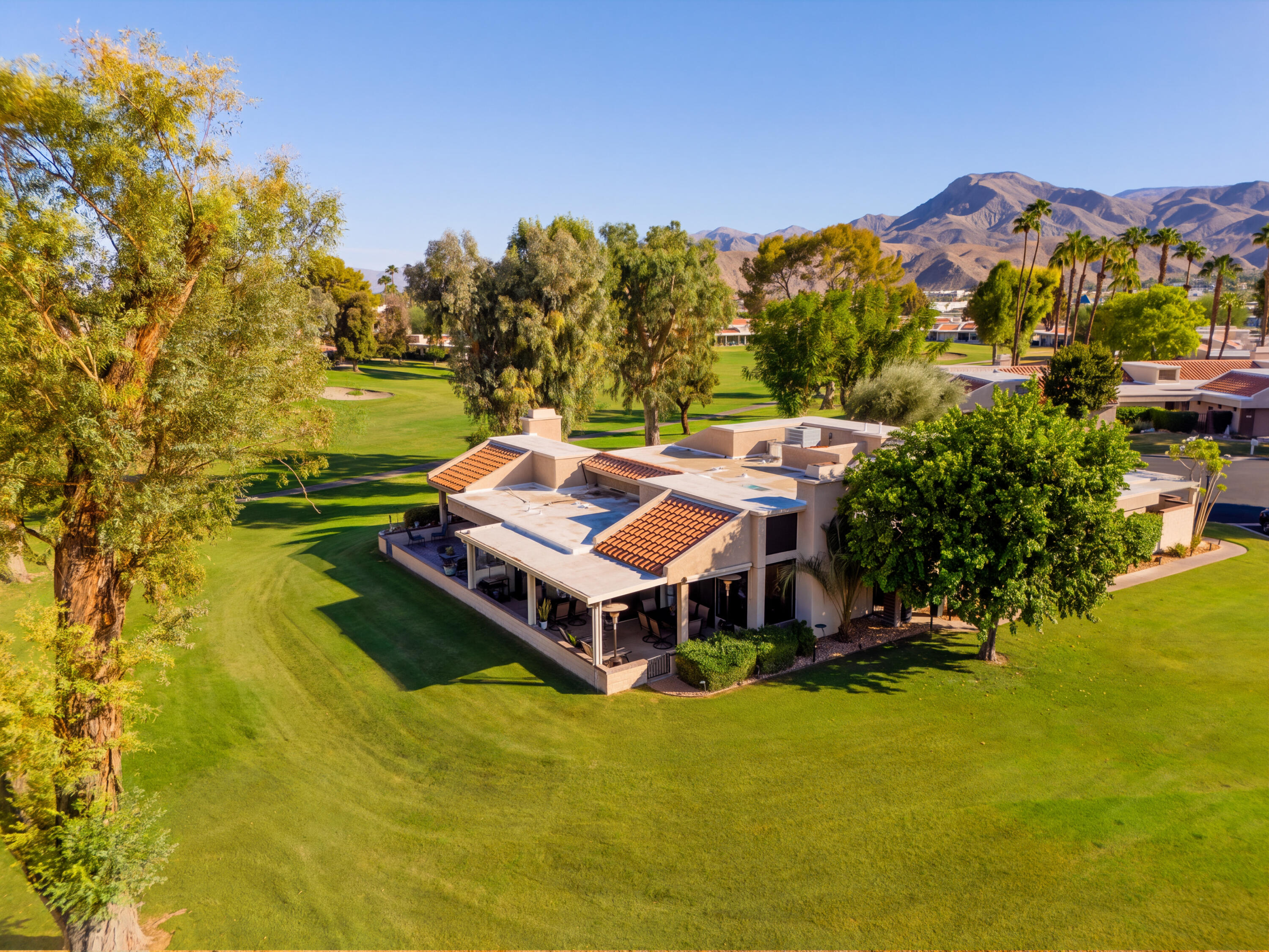 Aerial view of a tan home beside a golf course, with palm trees and desert mountains beyond.