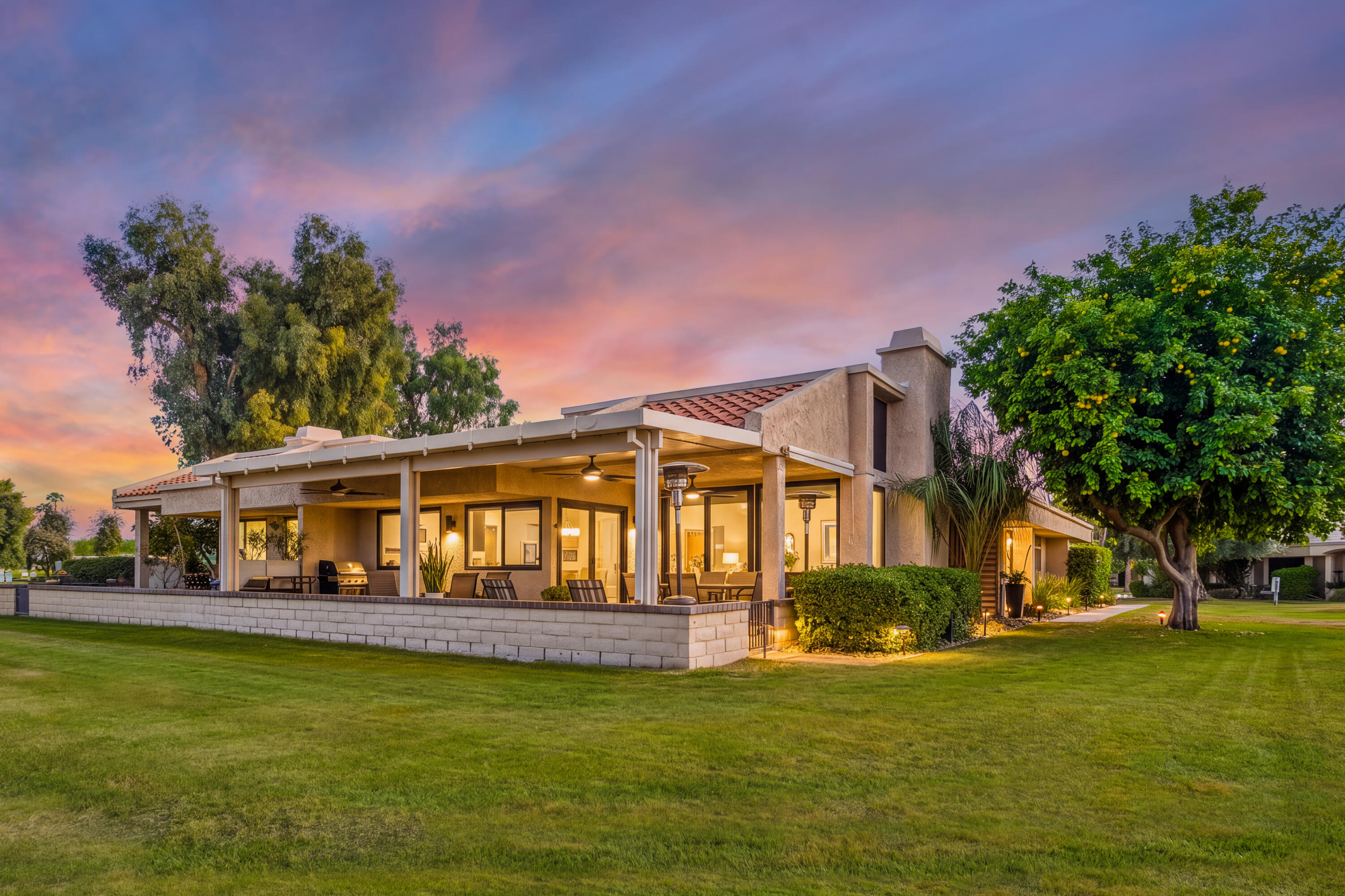 Stucco home with a large covered patio glowing at dusk, surrounded by a lawn and mature trees.