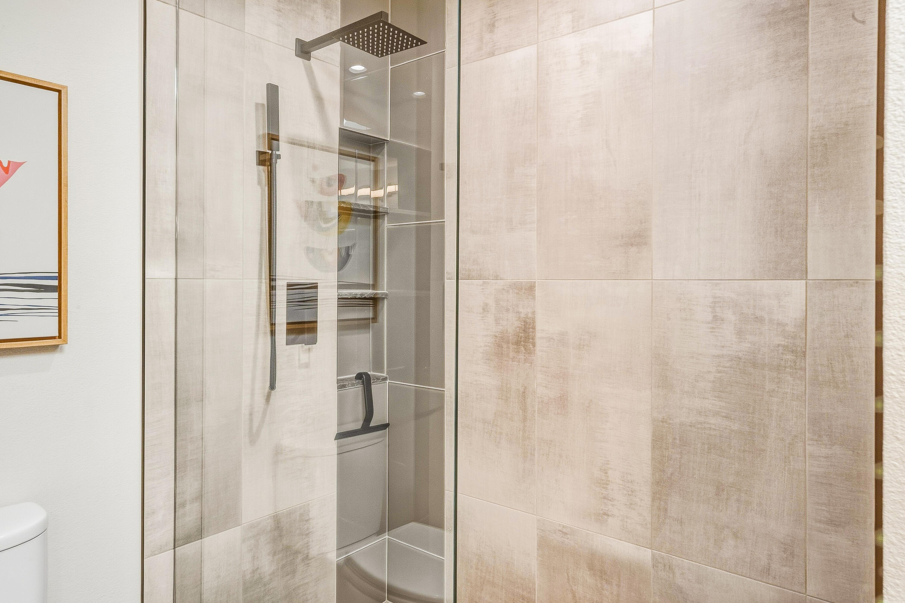 Bathroom shower with a glass enclosure, large beige tiles, rainfall showerhead, and black grab bar.
