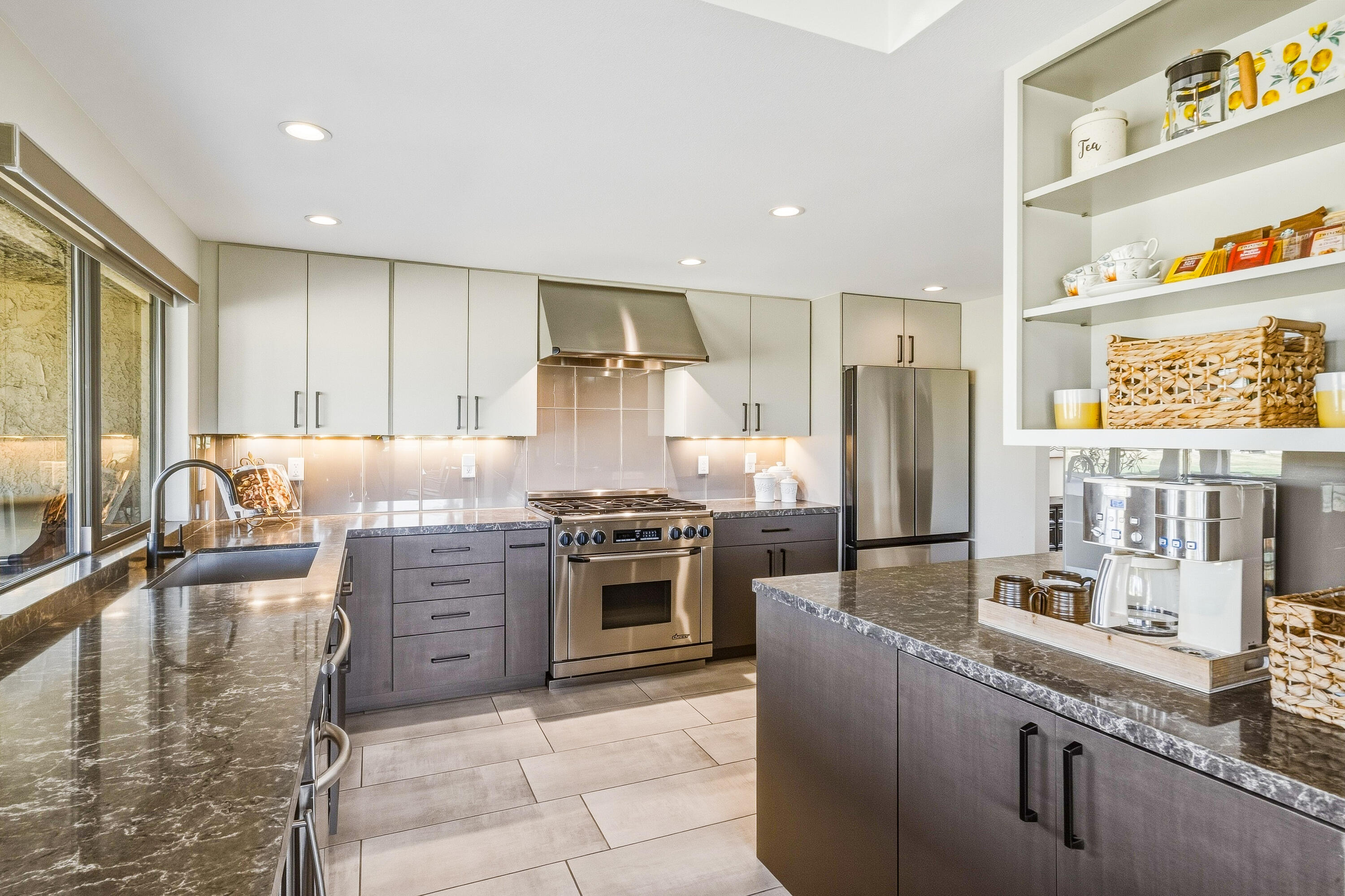 Modern kitchen with gray cabinets, dark stone counters, stainless steel appliances, and an island coffee station.