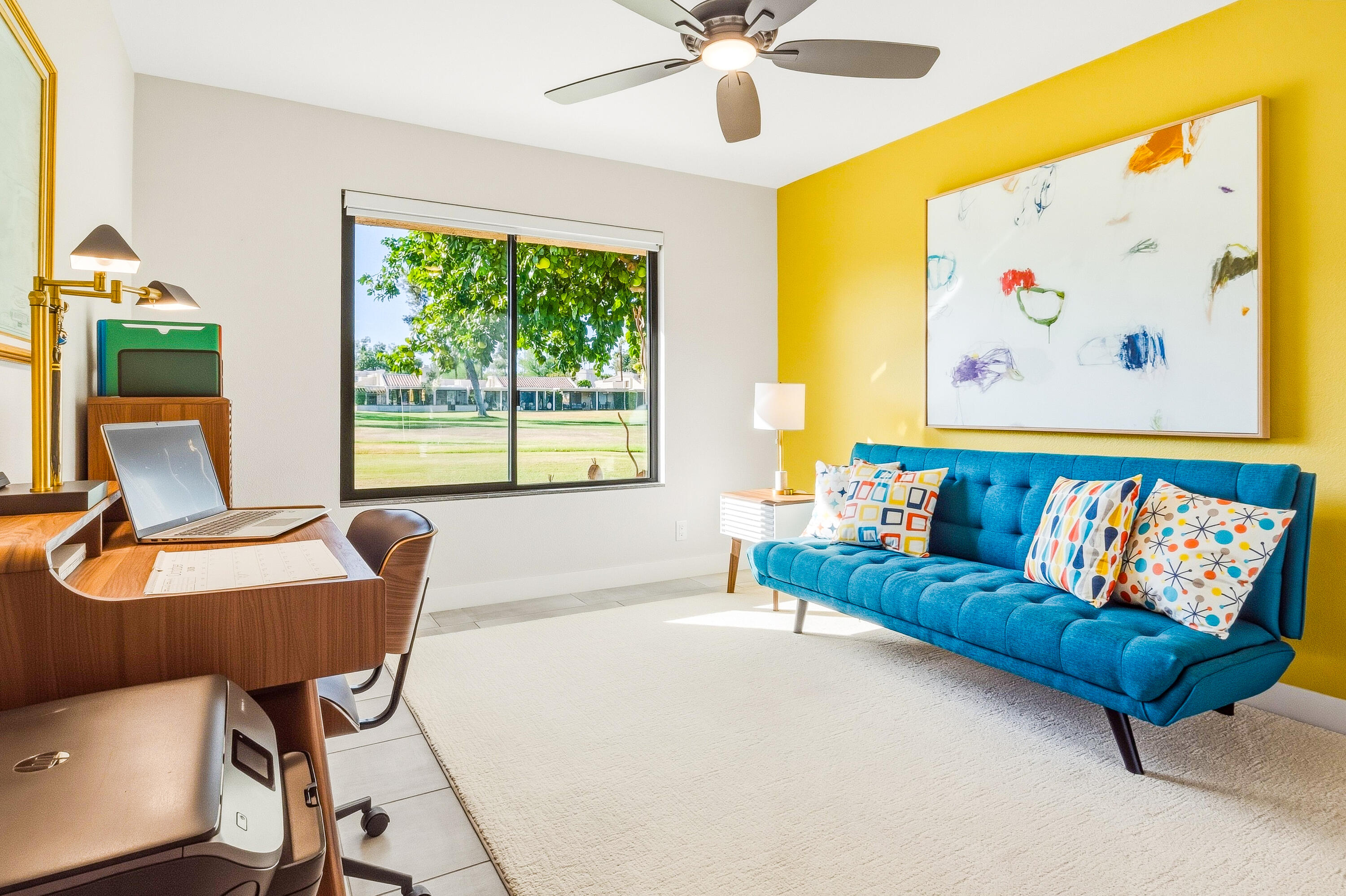 Living room with a blue sofa, yellow accent wall, and desk beside a window overlooking a lawn.