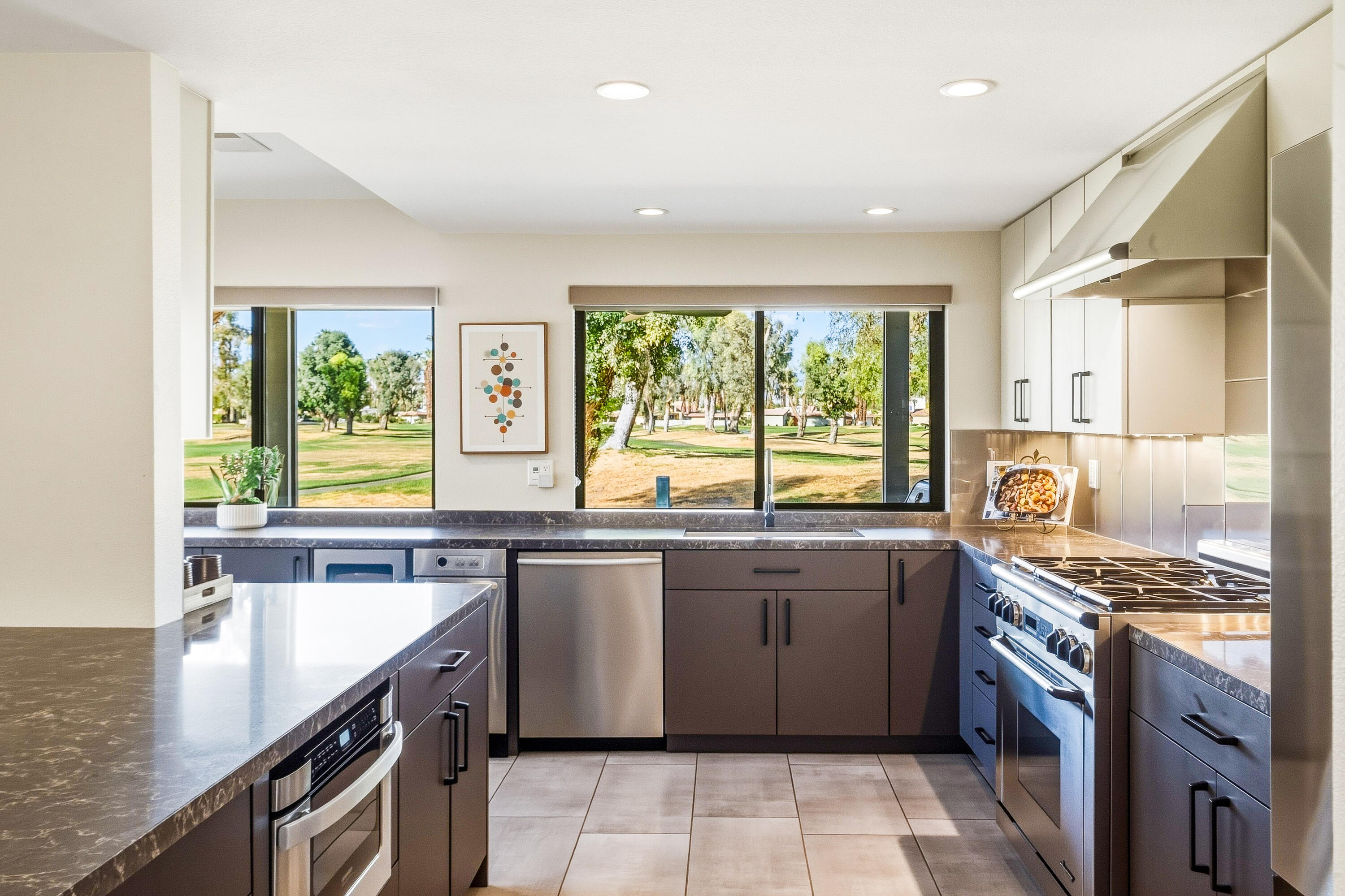 Modern kitchen with dark cabinets, stone counters, stainless appliances, and windows overlooking a green landscape.